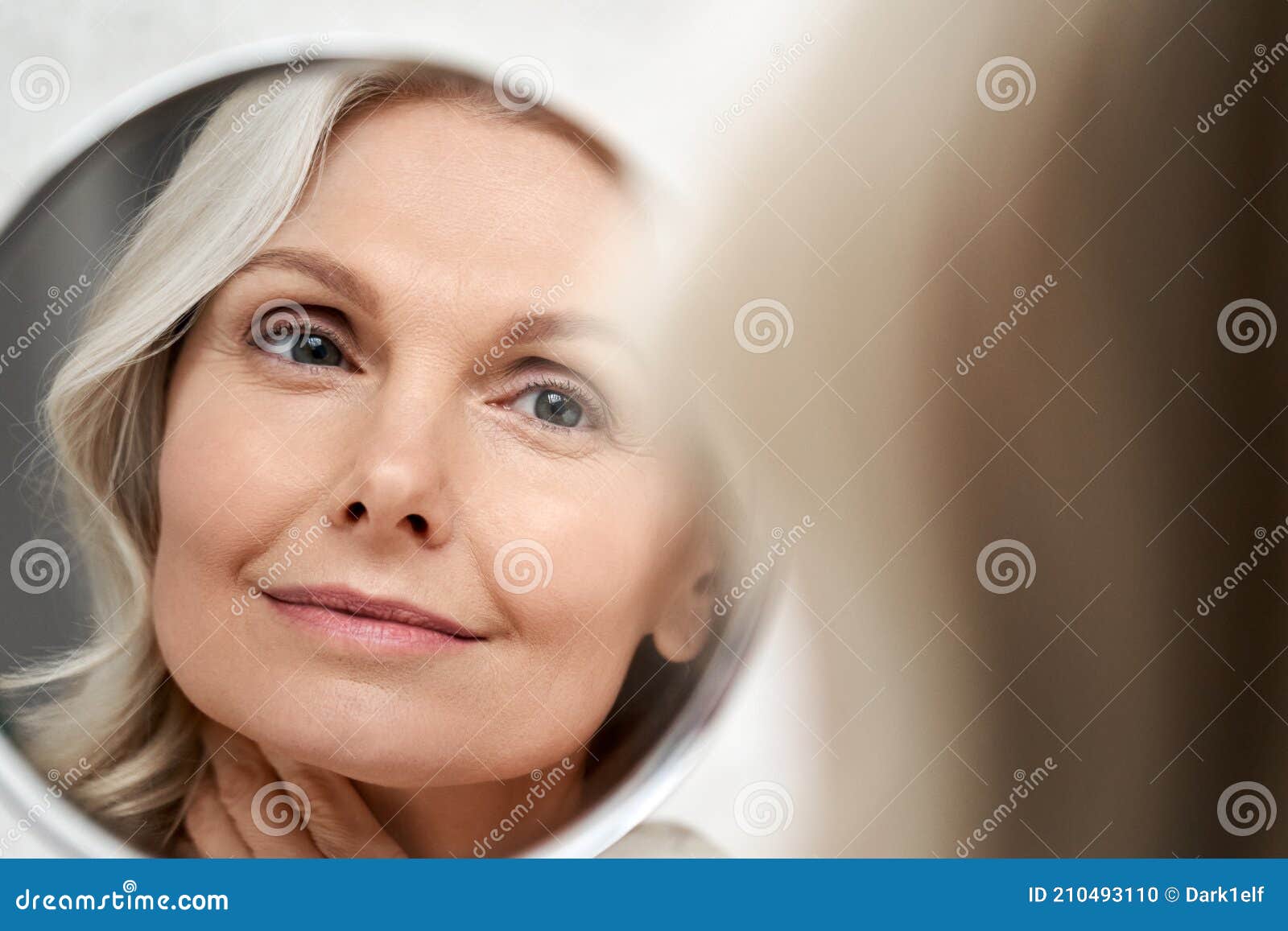 happy 50s middle aged woman touching face skin looking in mirror reflection.
