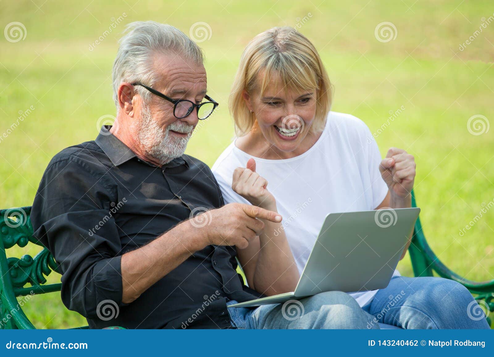 happy senior loving couple relaxing with laptop computer at park excited together in morning time. old people sitting on a bench