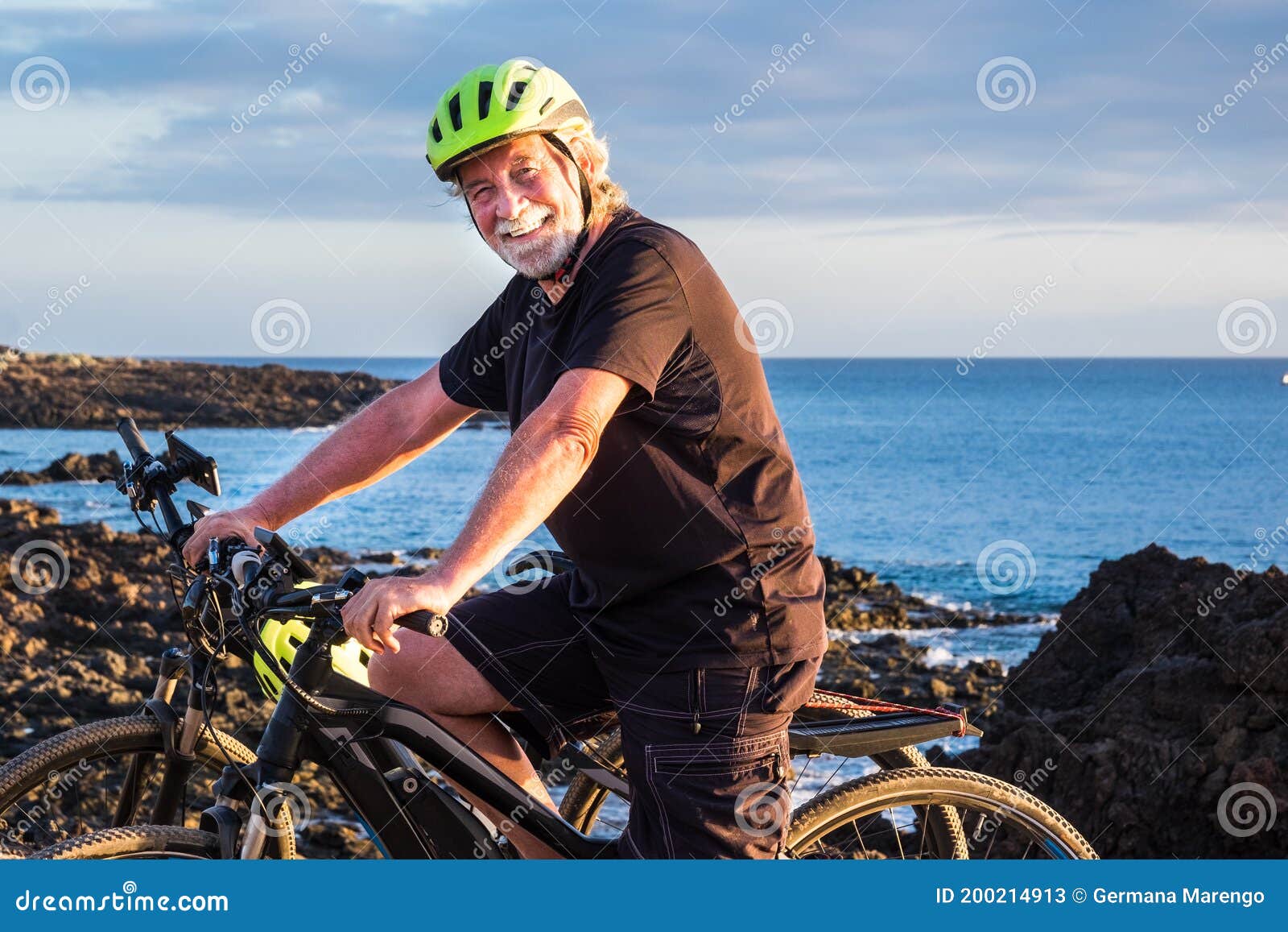 happy senior man standing on the cliff riding his bici and looking at camera.two electric bicycle close to him. blue ocean water