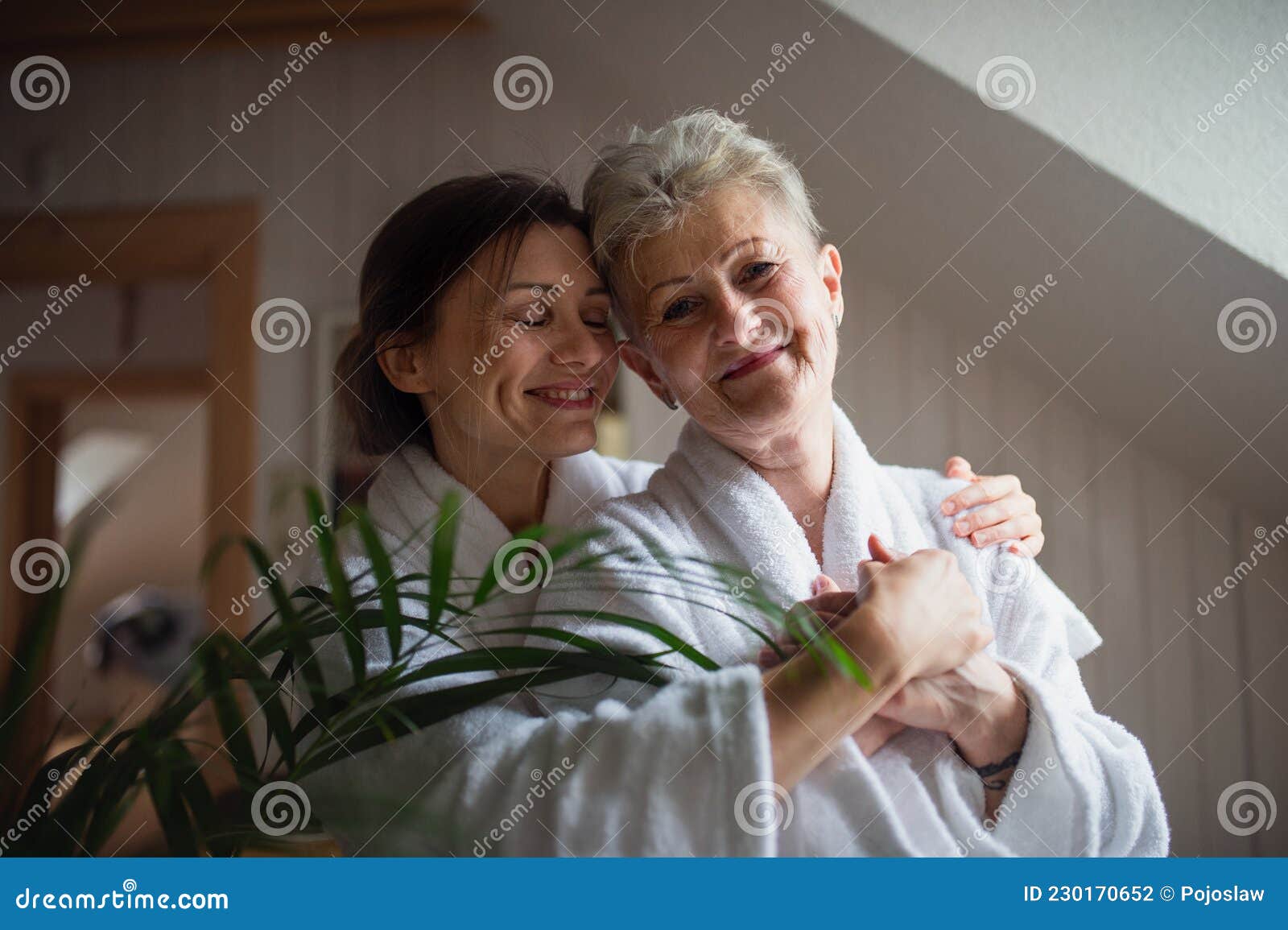 happy senior mother in bathrobe with adult daughter hugging indoors at home, selfcare concept.