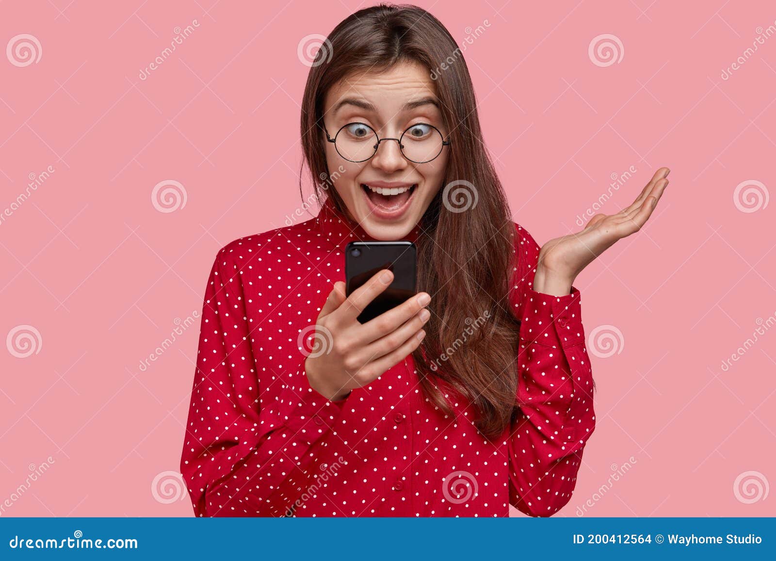happy surprised sociable lady looks positively at screen of cell phone, recieves good news, uses modern technologies
