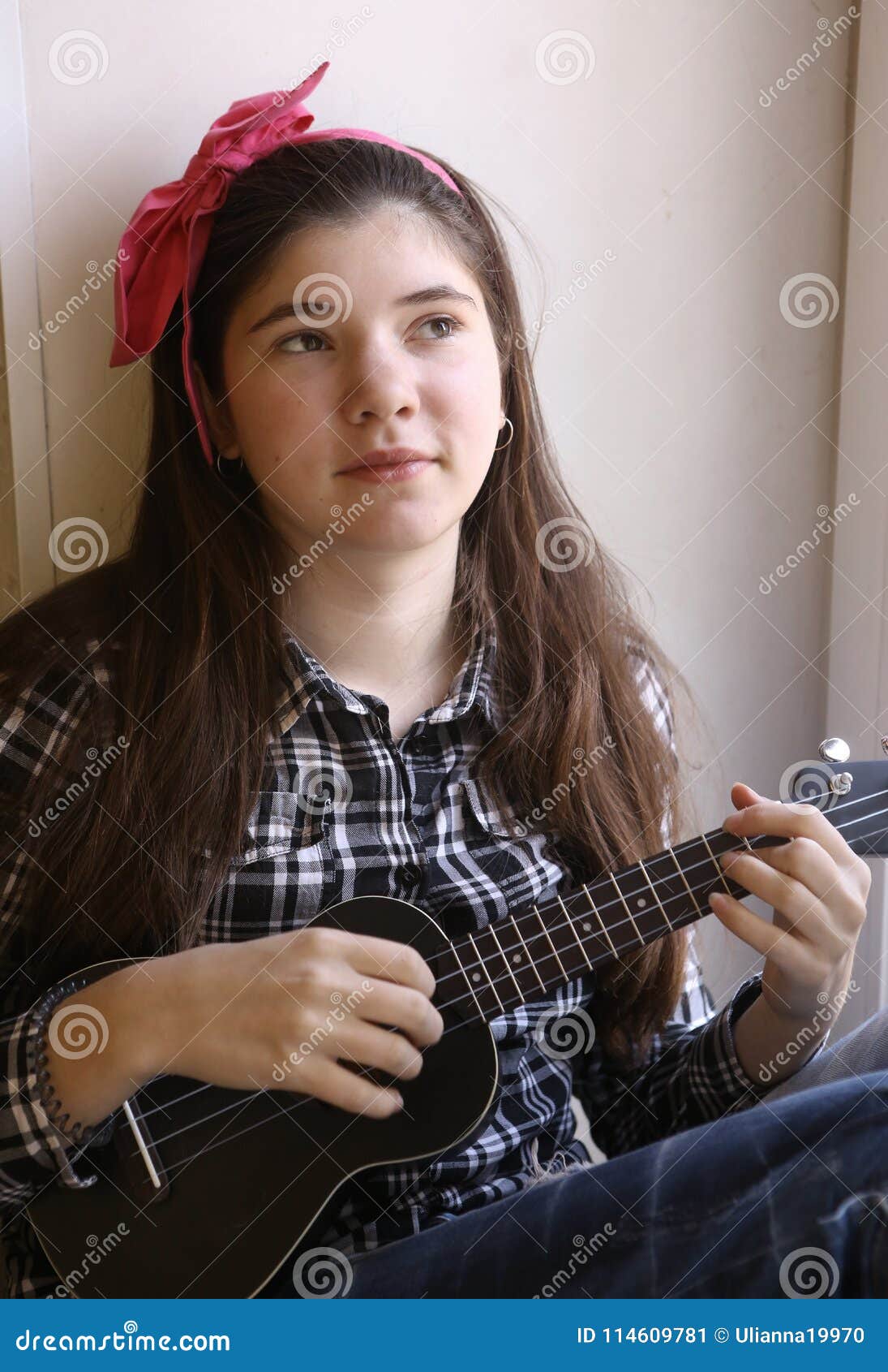 Smiling barefoot teen girl wearing lasses black leggings, grey shirt  playing song on yellow ukulele guitar while sitting on wooden stairs at  home indoors, looking at camera. Music and hobbies concept Stock