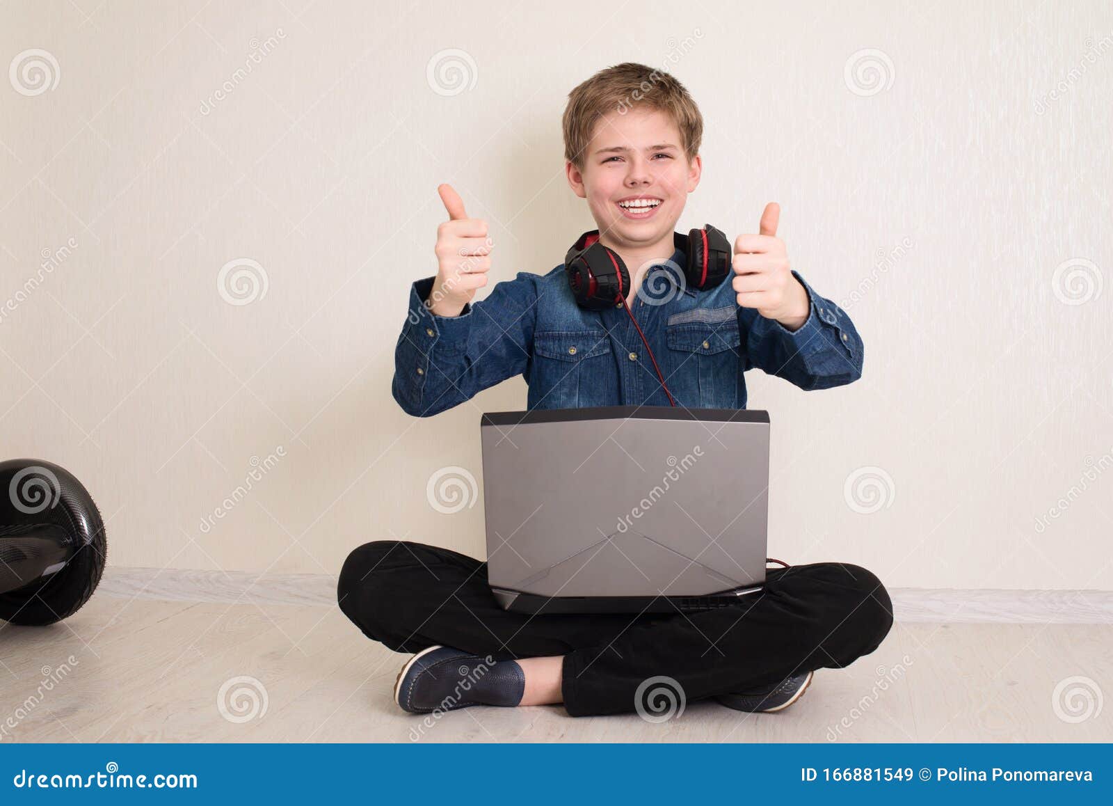 Happy Teenager Junge mit Laptop-Computer und Kopfhörer mit Daumen auf der Geste, während er auf dem Boden sitzt, die Beine gekreuzt