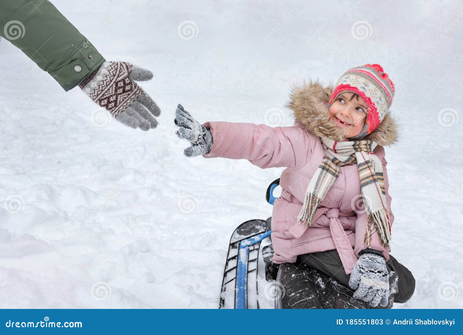 a happy toothless girl is sitting on a sledge in a winter park, reaching for an adultÃ¢â¬â¢s hand before touching