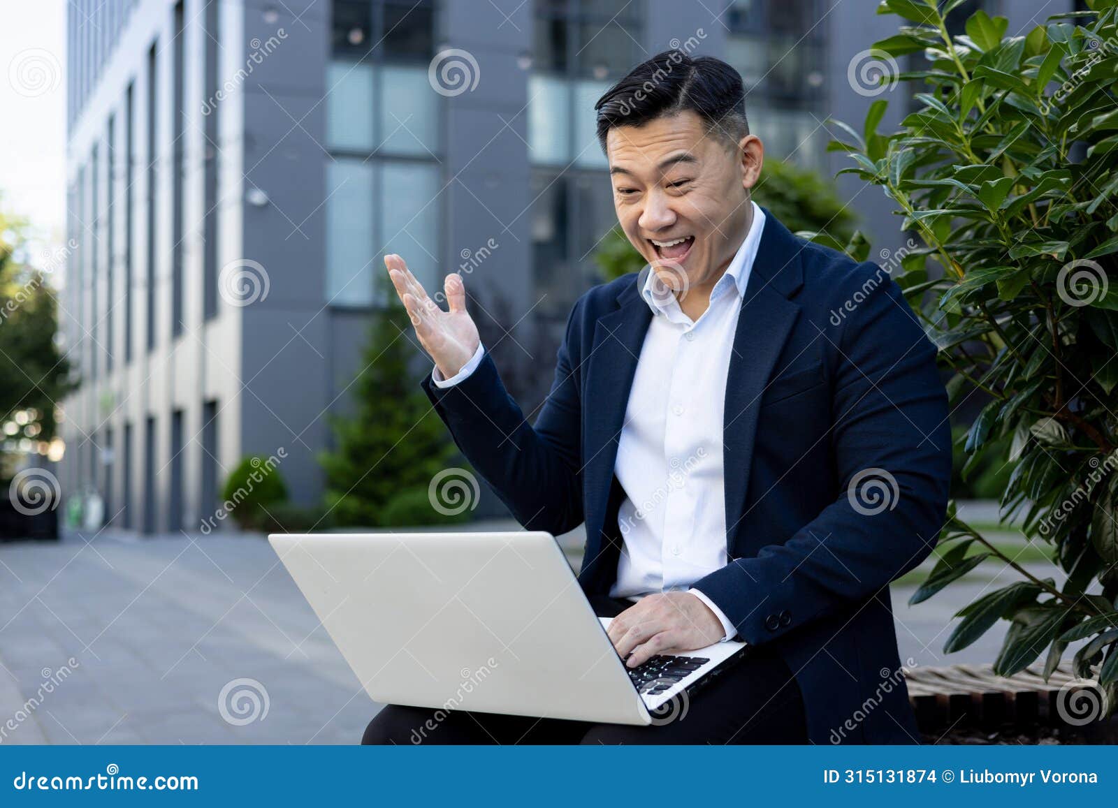 a happy young asian male businessman is sitting outside an office building and smilingly looking at the screen of a