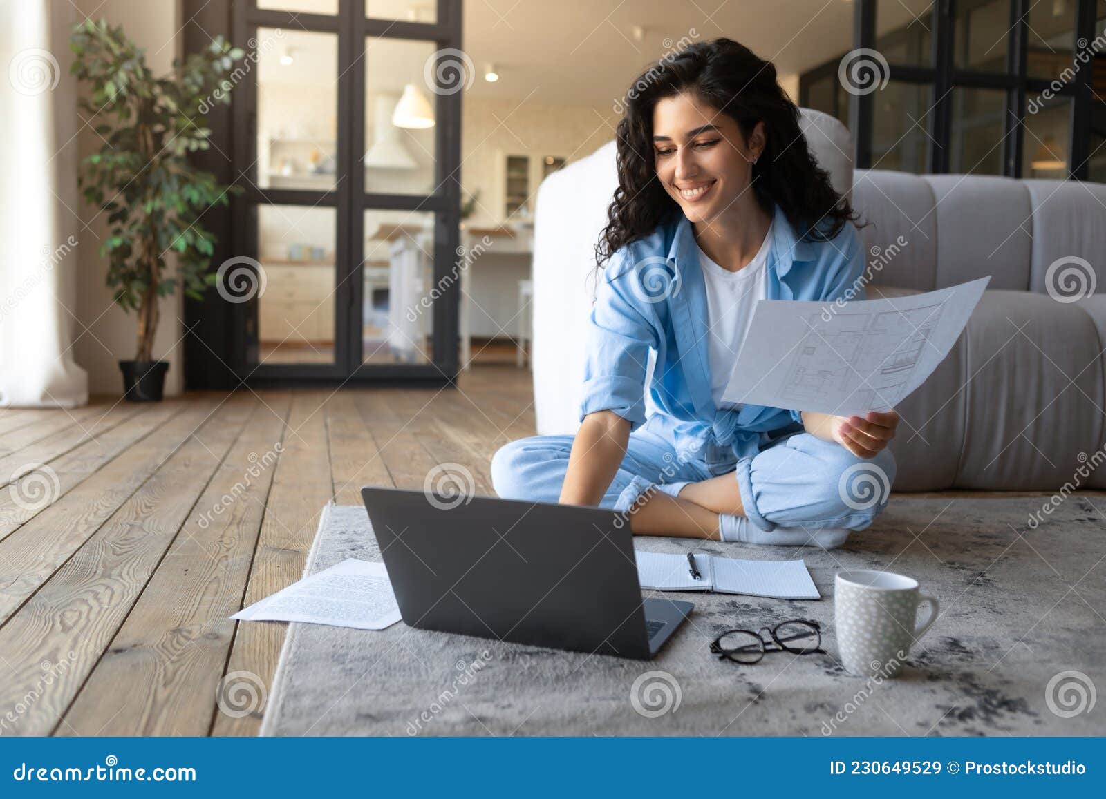 happy young cauasian lady working or studying on laptop computer, holding documents, looking at pc screen indoors