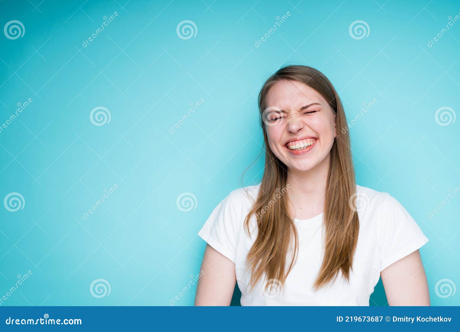 happy young girl in a white t-shirt smiles broadly showing teeth and closes her eyes while standing on a blue background