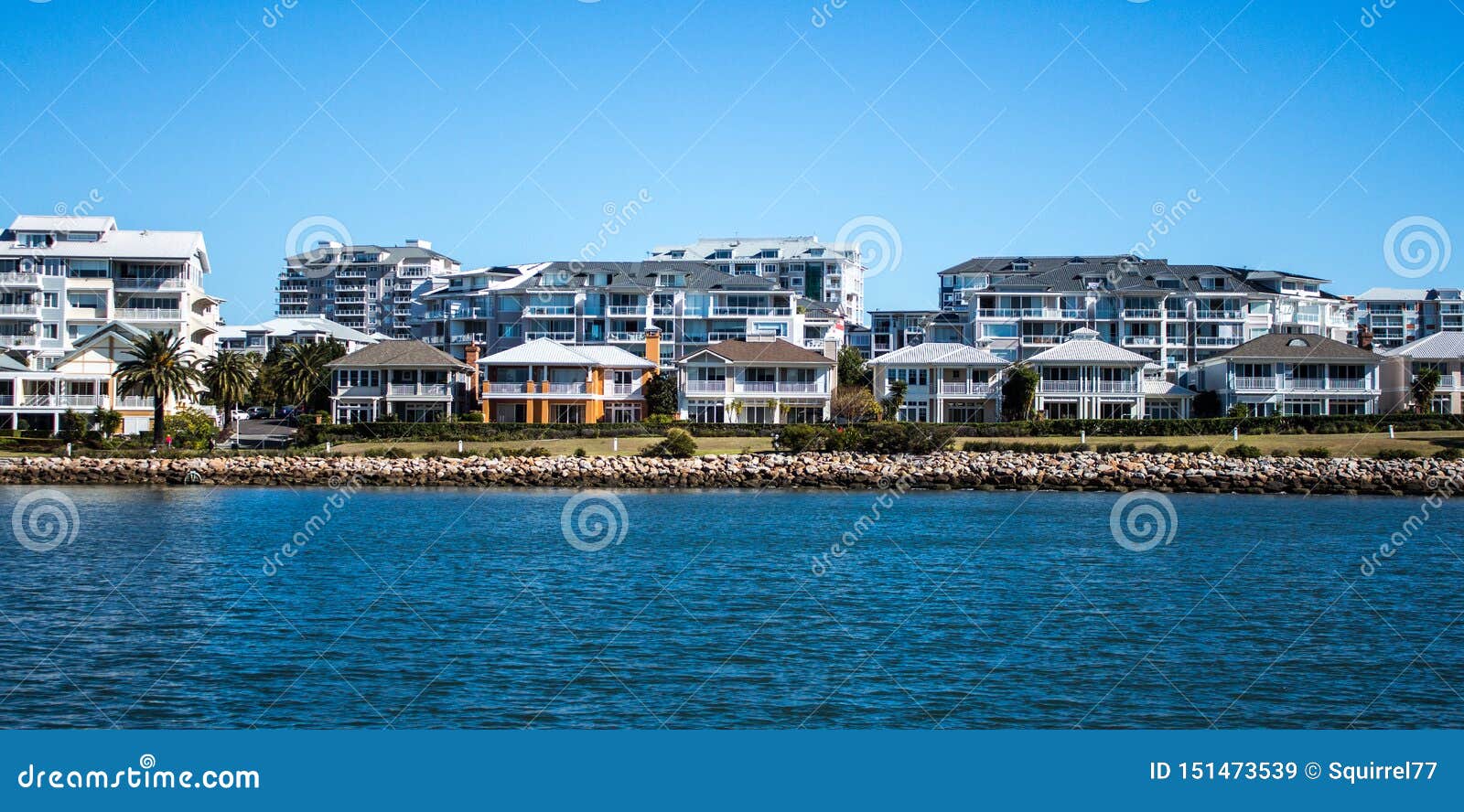 harbourside condominium apartments behind free standing houses with grass frontage, stone retaining wall and blue river against bl
