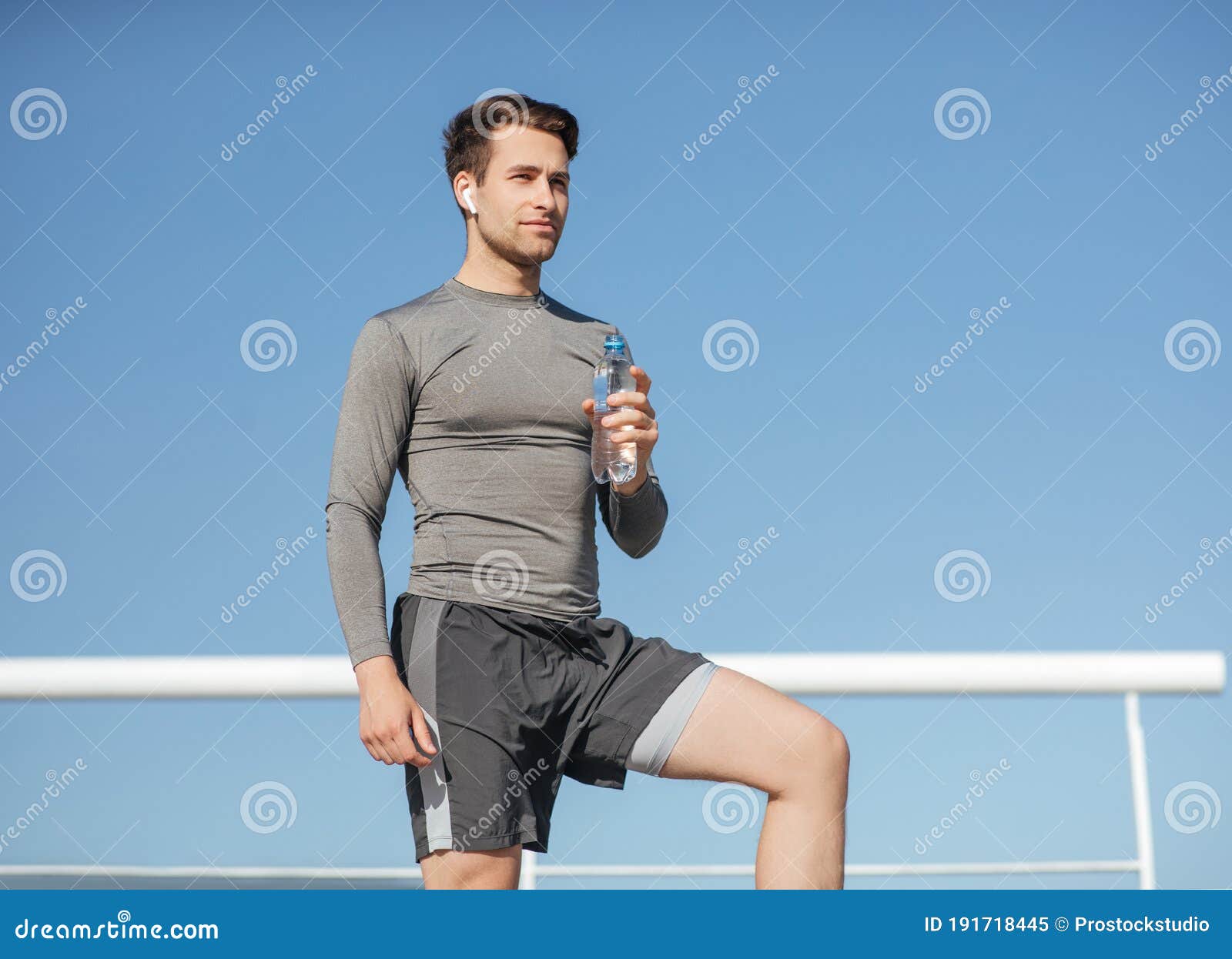 health and body care. muscular handsome guy in sportswear, with wireless headphones holding bottle of water on blue sky