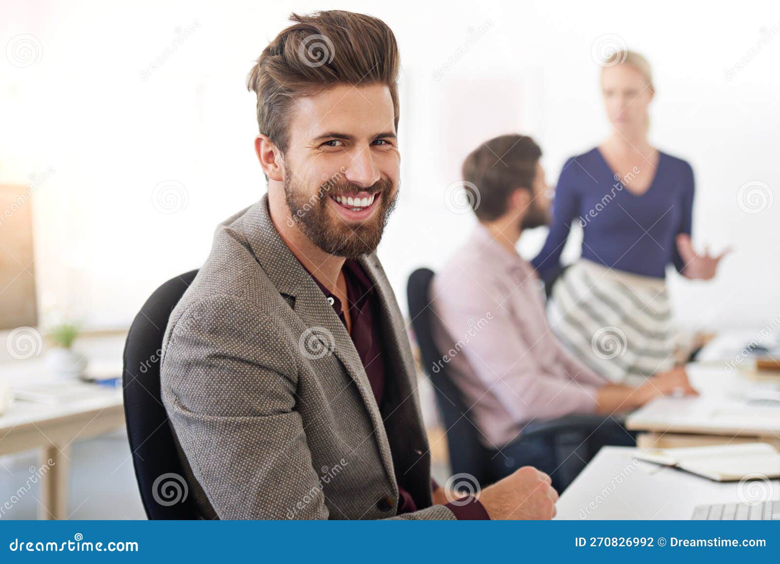 hes one confident exec. a businessman sitting at his desk with his coworkers in the background.