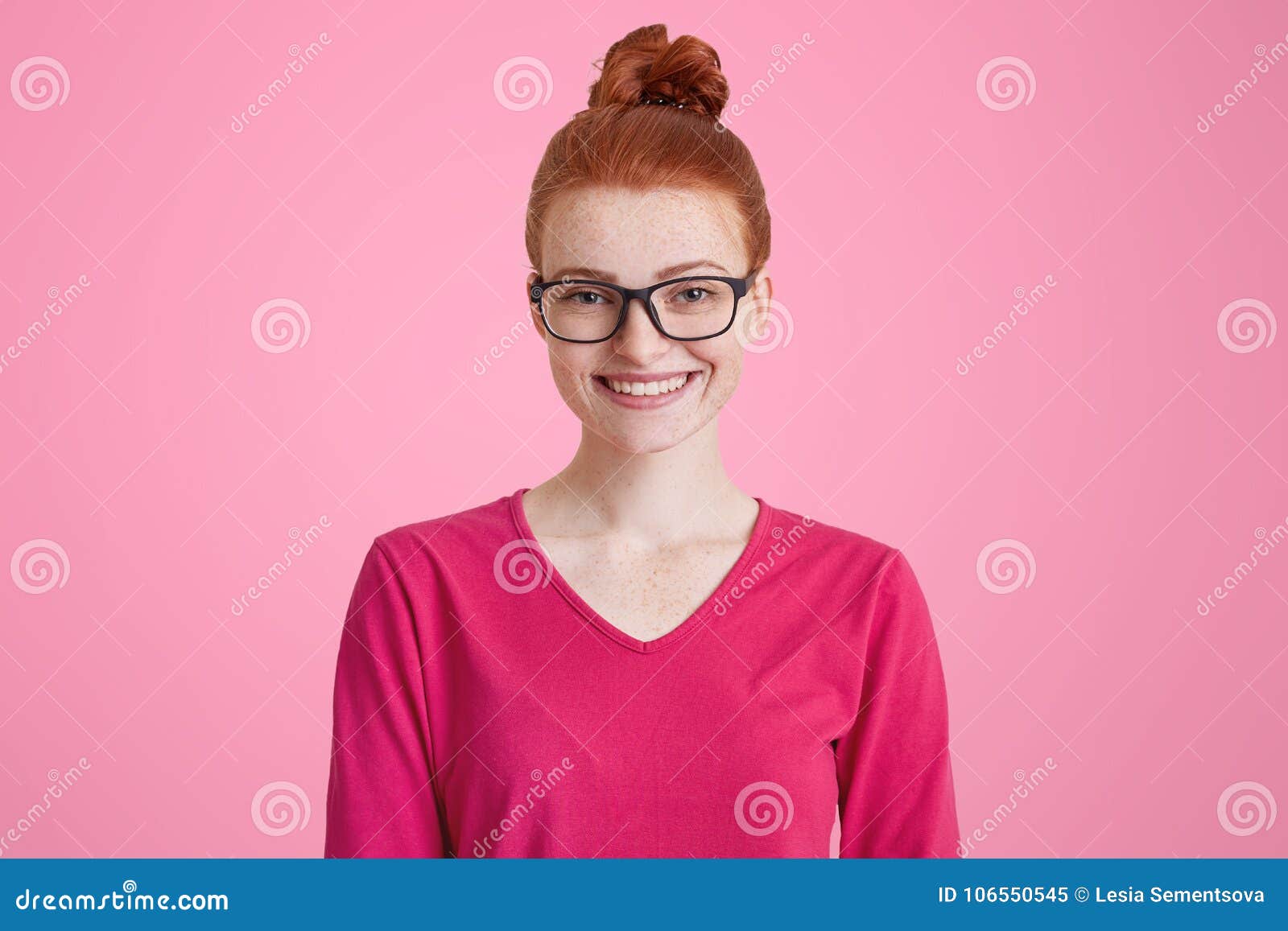 Het geïsoleerde schot van gelukkige gembervrouw in roze sweater, draagt vierkante bril, heeft rood die haar in knoop, freckled huid, prettige die glimlach wordt gebonden, over roze achtergrond wordt geïsoleerd Vrolijke vrouwelijke student binnen