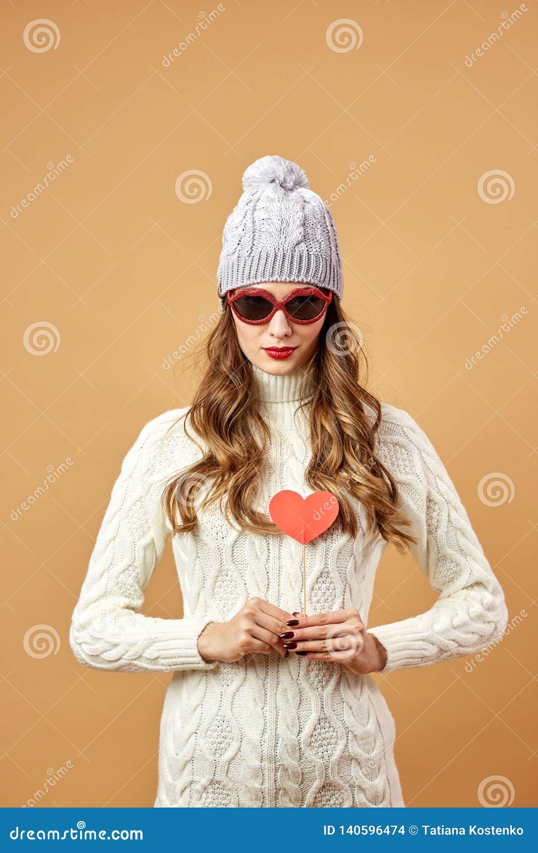 Het grappige meisje in zonnebril kleedde zich in witte gebreide sweater en hoedenrijkenpret met een rood document hart op een stok op een beige achtergrond in de studio