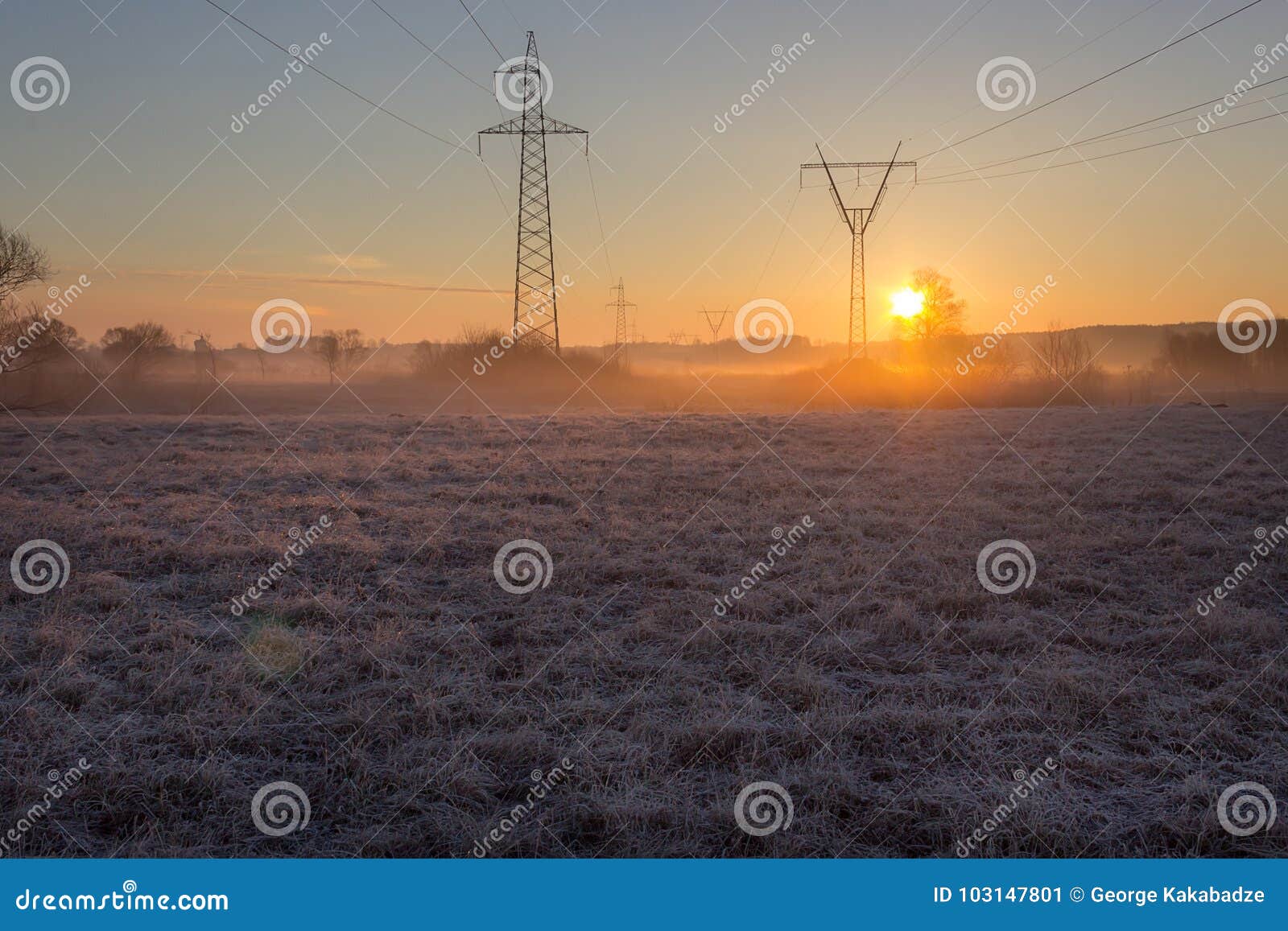 High voltage line supports in fog at frosty spring sunrise morning. High voltage line supports at early morning in frosty field with old grass shining at sunrise