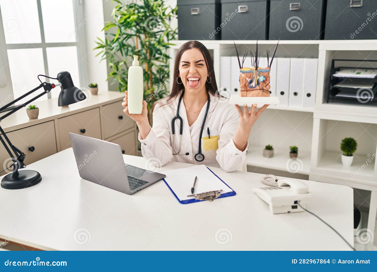 hispanic doctor woman holding model of human anatomical skin and hair sticking tongue out happy with funny expression