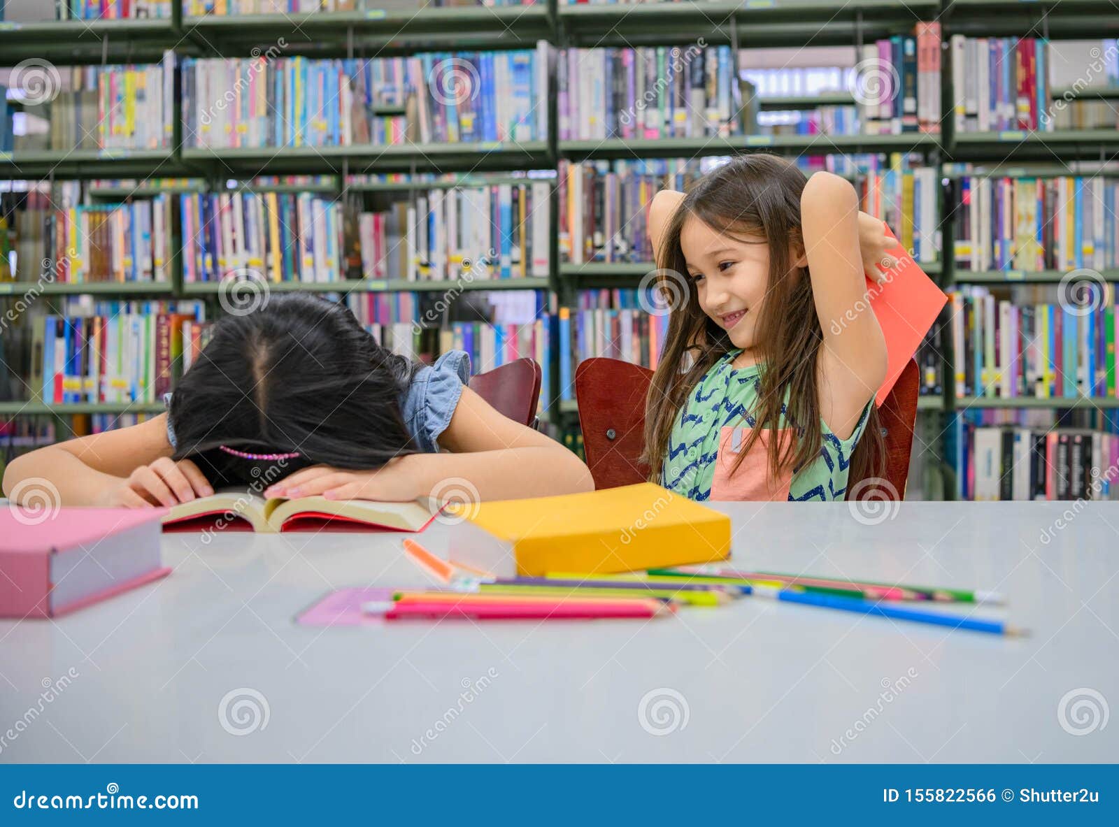 HjÃ¤rtliga flickor som lÃ¤ser bok och leker med att slÃ¥ en sovande vÃ¤n i skolbiblioteket. MÃ¤nniskors livsstil och utbildning och vÃ¤nskap. Fritidsaktiviteter fÃ¶r barn- och barngrupp