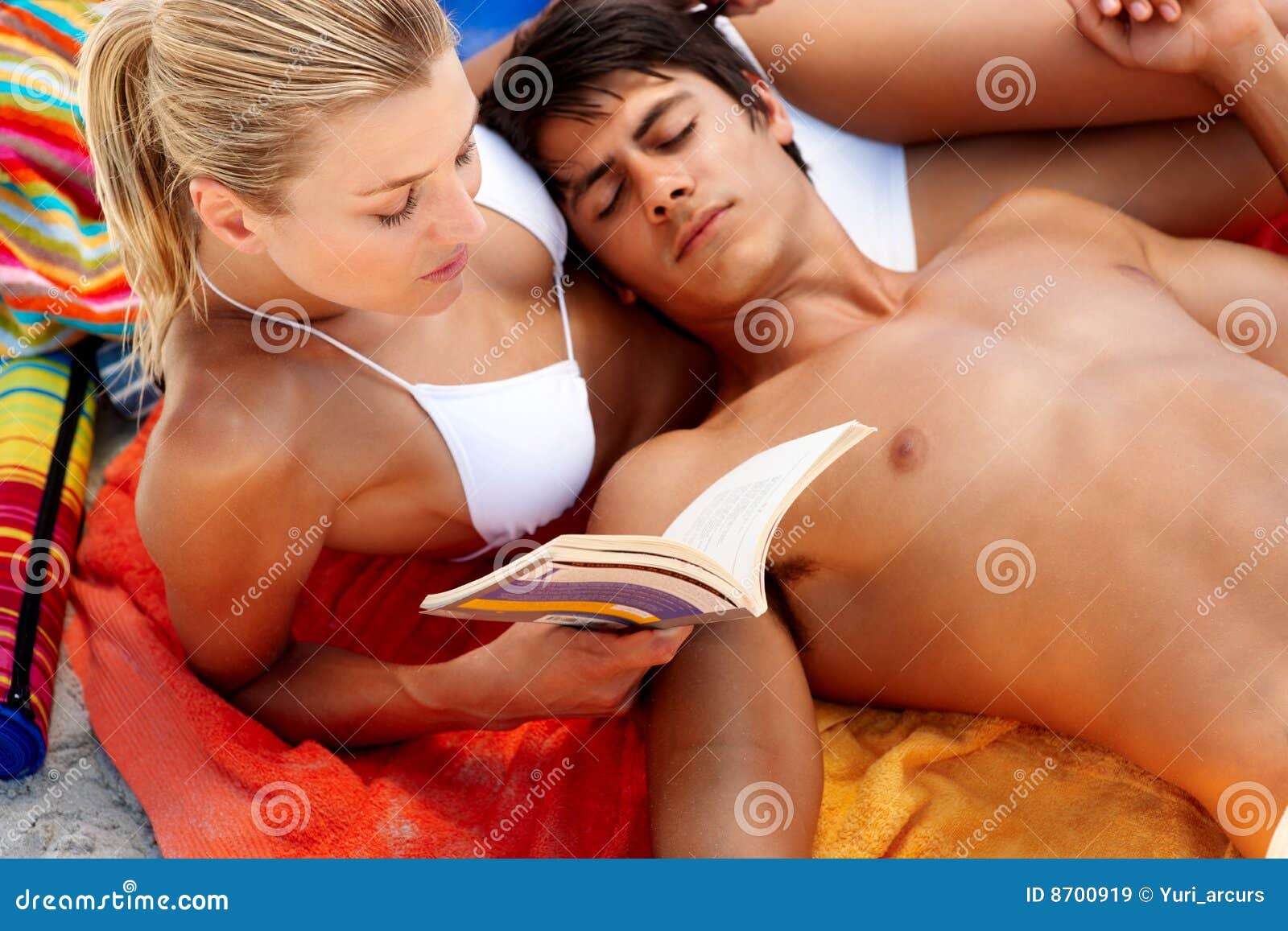 Hombre joven que se reclina mientras que su novia está leyendo. Mujer joven que lee un libro y a un hombre que duermen en ella en la playa