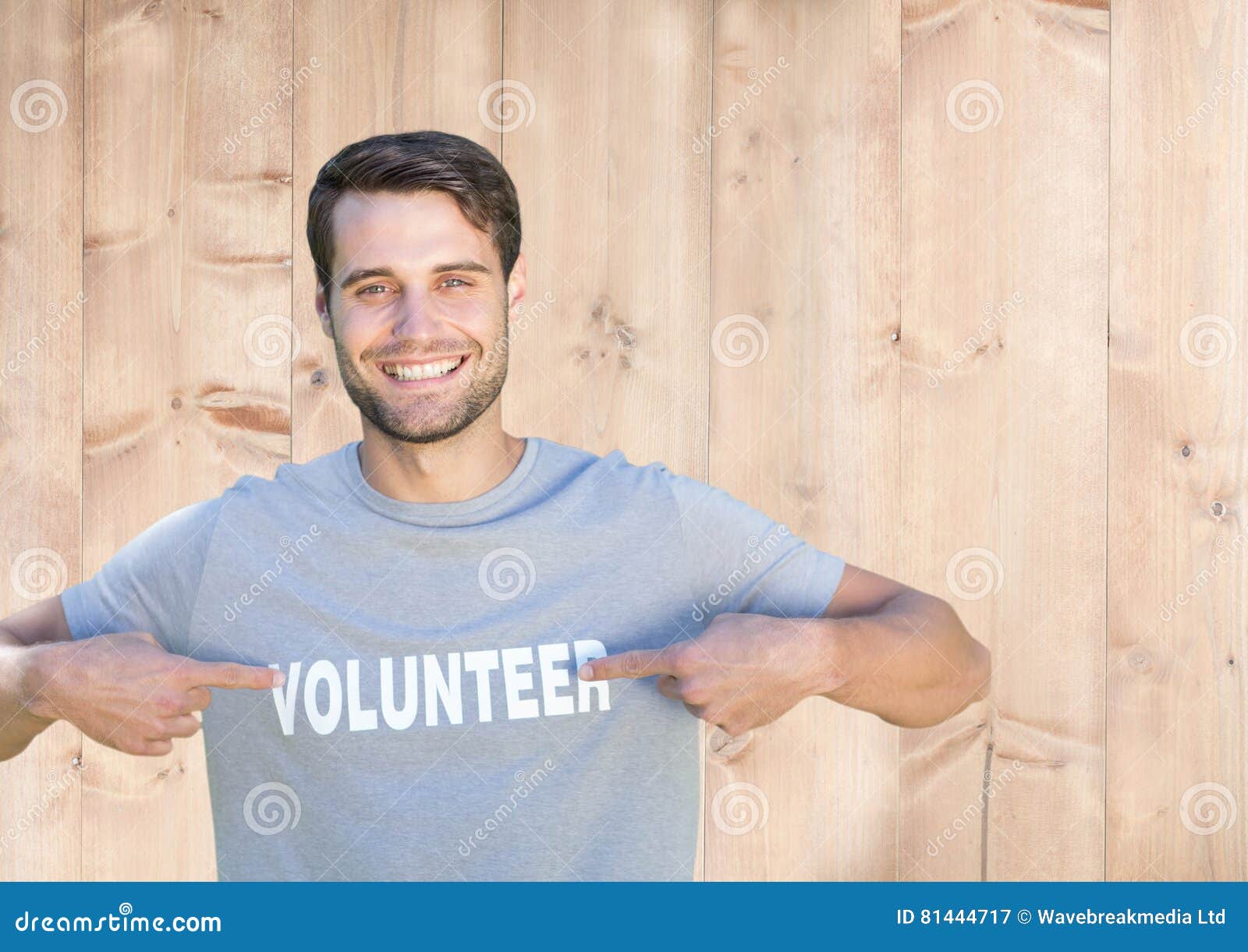 Hombre sonriente que señala en el título voluntario en su camiseta. Retrato de los hombres sonrientes que señalan en el título voluntario en su camiseta contra fondo de madera