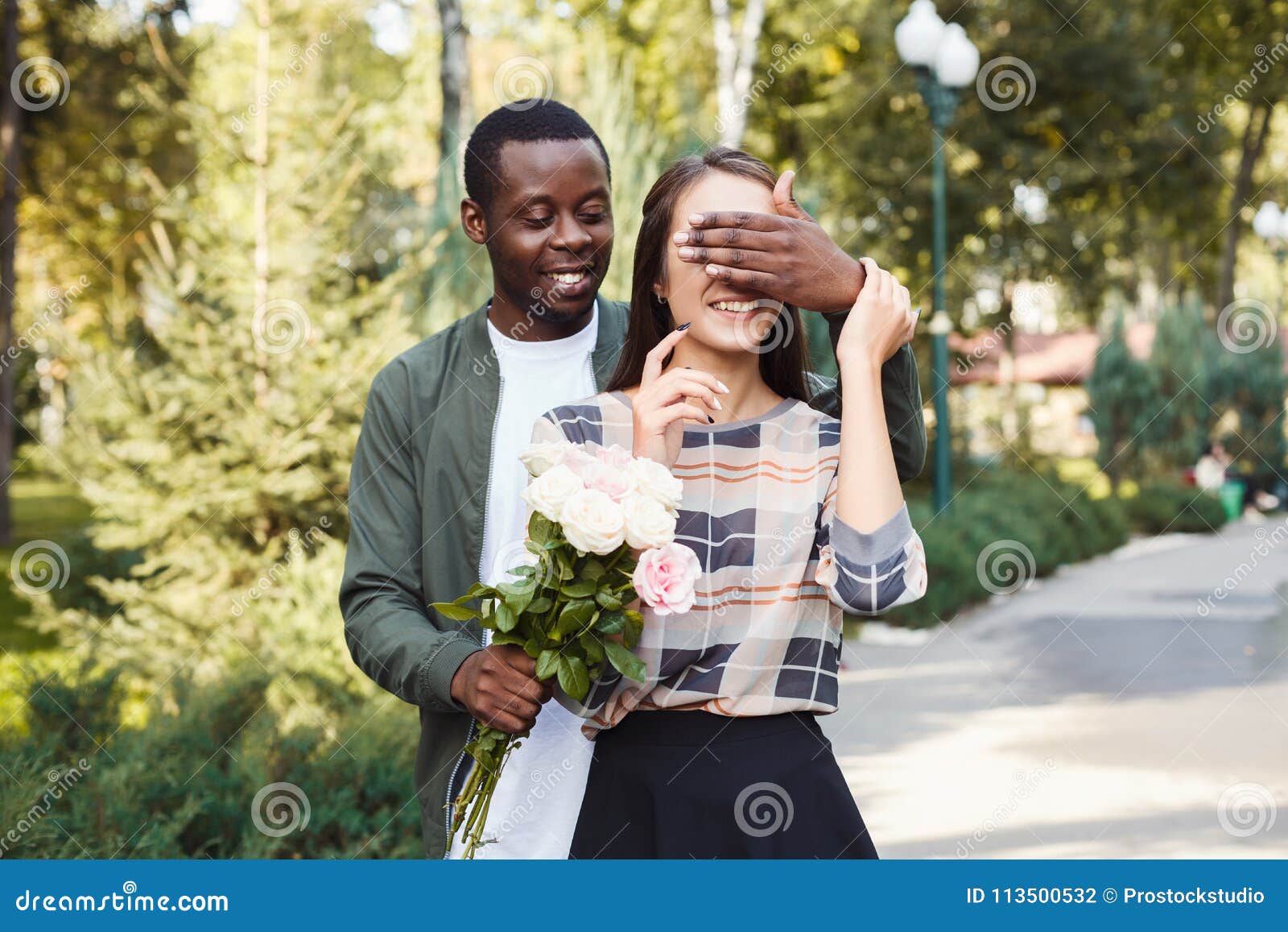 Homem surprising sua amiga com flores. Os homens afro-americanos novos com as flores que fecham sua amiga eyes para fazer a surpresa para ela, andando no parque