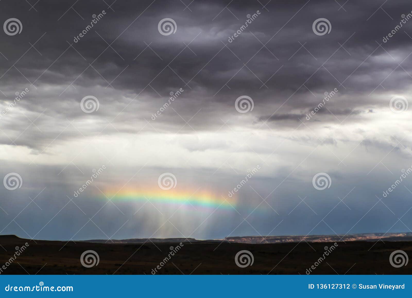 horizontal rainbow across the middle of a rain squall near the horizon in desert with distant cliffs lit up and foreground dark -