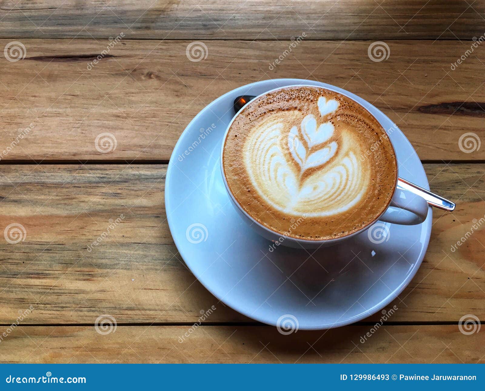 hot cappuccino coffee in white cup and saucer with spoon on wooden table background. art of milk foam drawing.