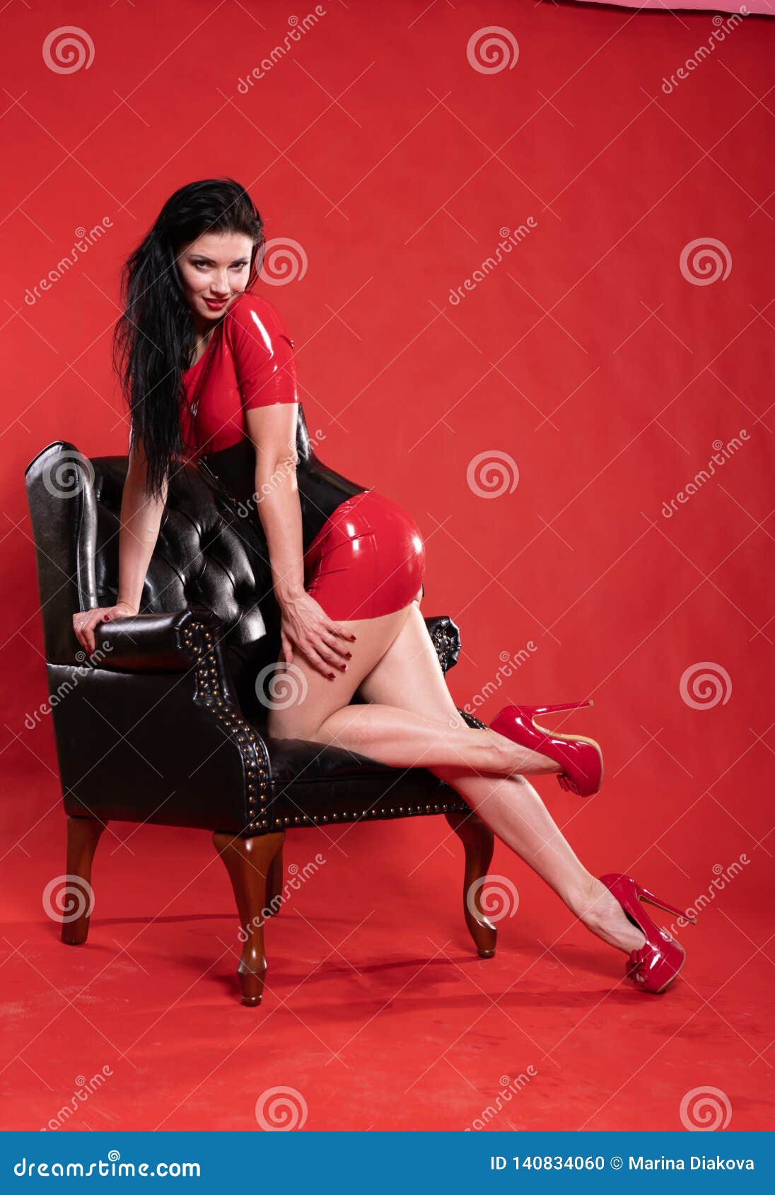 Hot Girl Wearing Latex Rubber Outfit and Posing with Leather Chair on Red Studio Background Alone Stock Photo