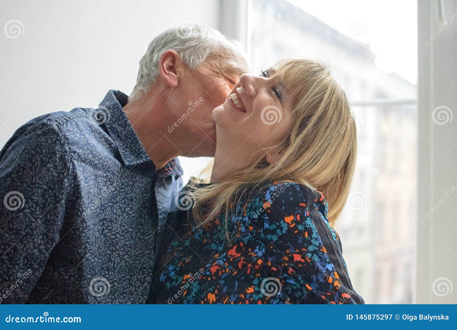 Hot and Middle-aged Woman Enjoying Hug of Her Elderly Husband Standing Near Opened Window Inside Their Home