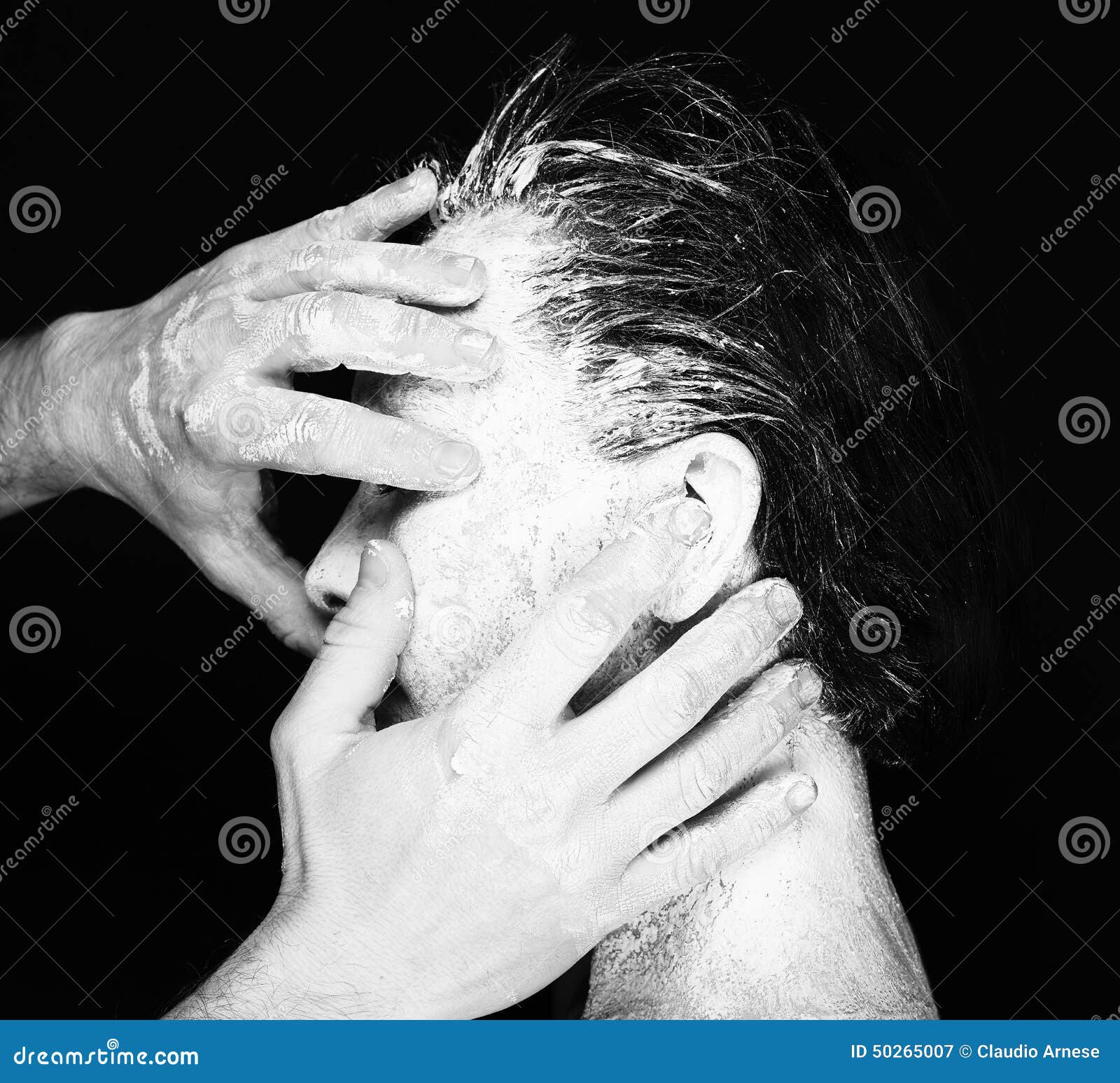 Black and white portrait of human hands working with woman with clay on face on black background