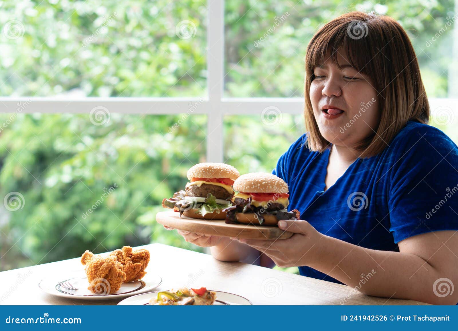 Hungry Overweight Woman Holding Hamburger On A Wooden Plate Fried Chicken And Pizza On Table