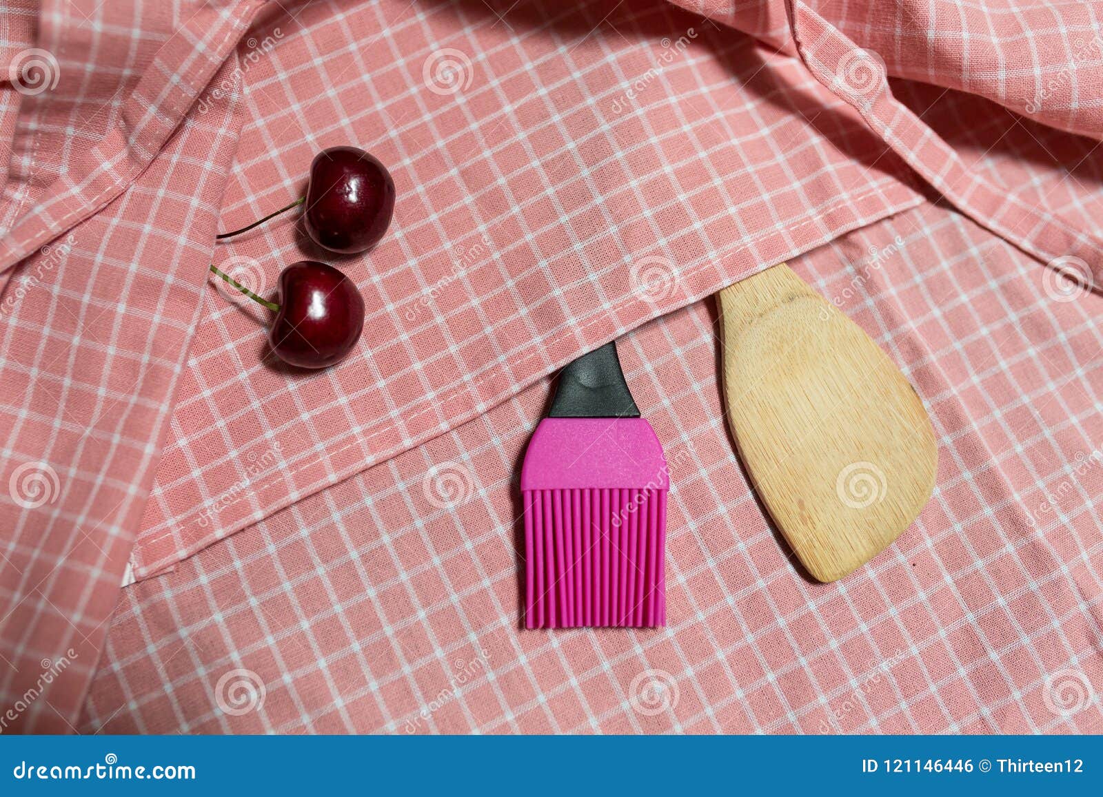 Hölzerne Schöpflöffel- und Silikonbürste auf rosa Gitter Stoff Mit Kirsche zwei. Hölzerne Schöpflöffel- und Silikonbürste in rosa Taschengitter Stoff Mit Kirsche zwei