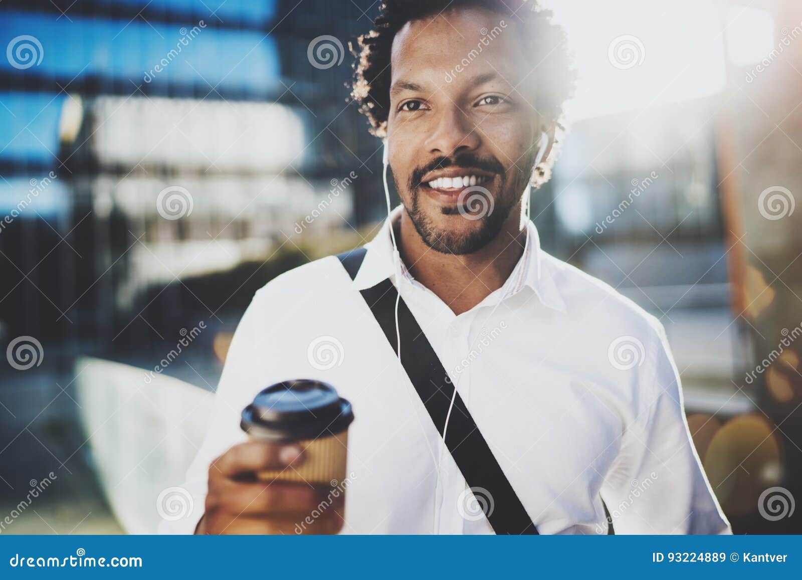 Il ritratto dell'uomo africano americano sorridente in cuffia che cammina alle vie soleggiate con caffè dentro porta via la tazza e godere di ascoltare musica sul suo smartphone Priorità bassa vaga