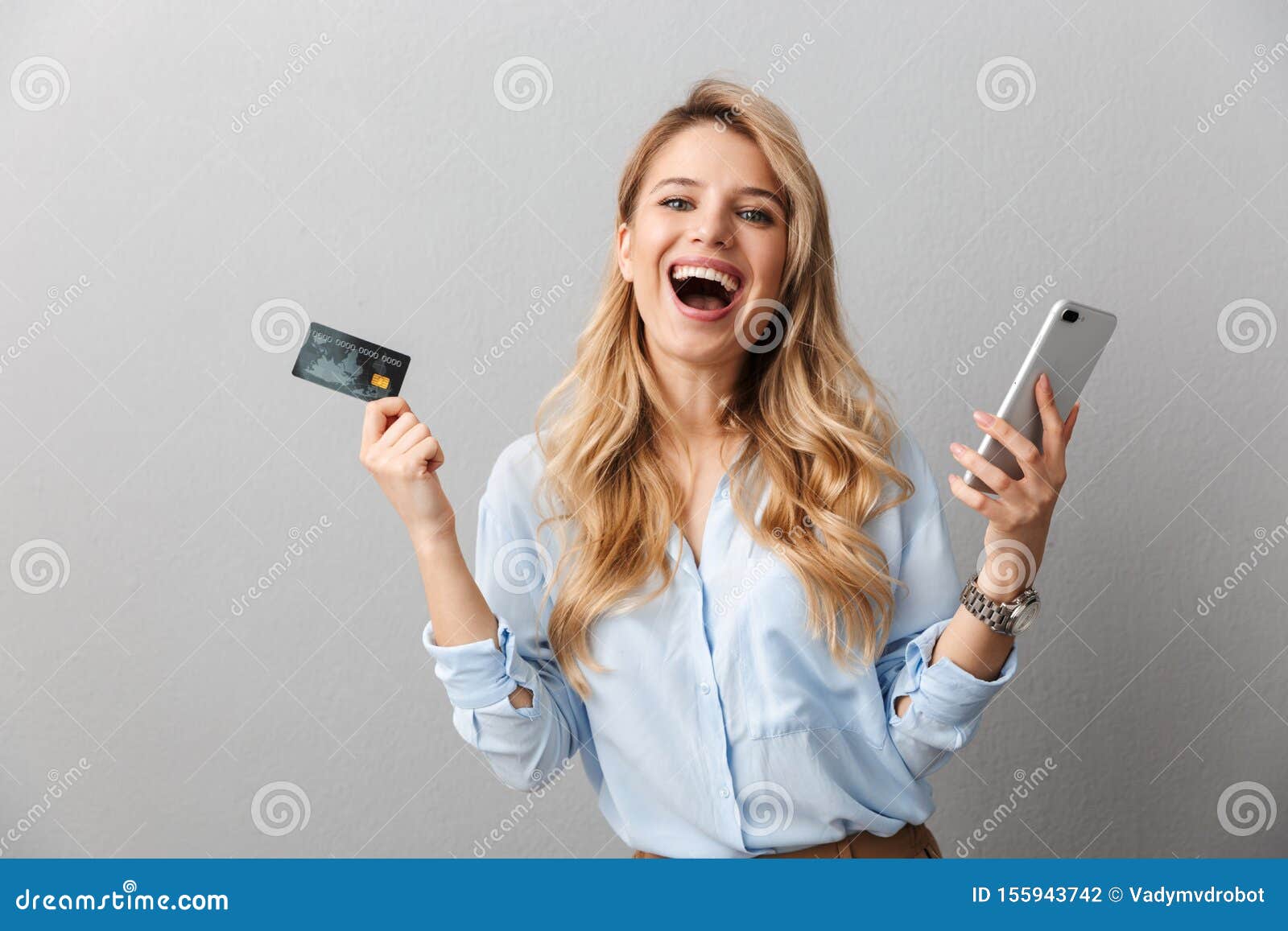 happy young pretty blonde business woman posing  grey wall background holding credit card using mobile phone