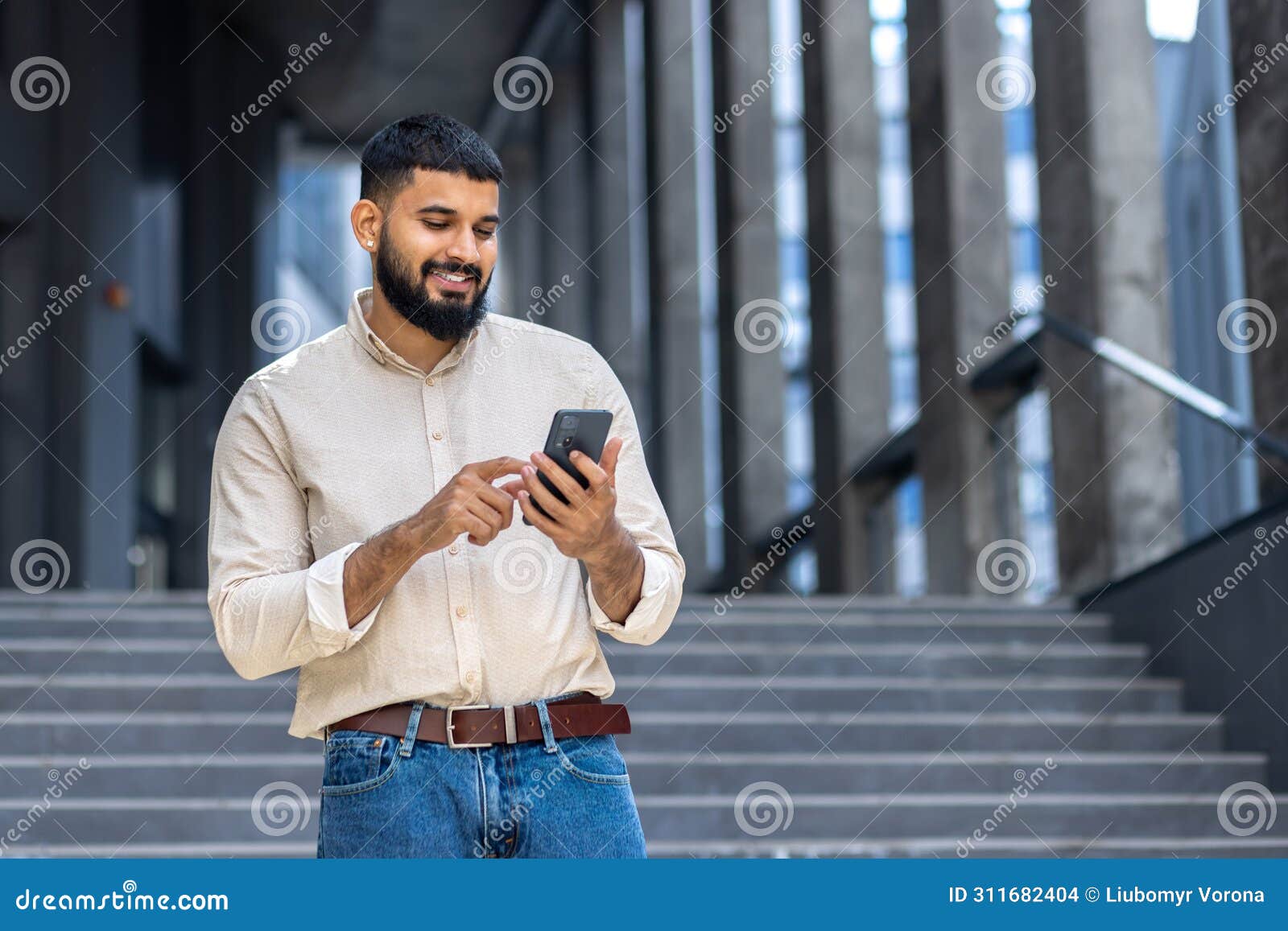 indian young male student standing on campus on stairs near building wearing shirt and jeans and smilingly using mobile