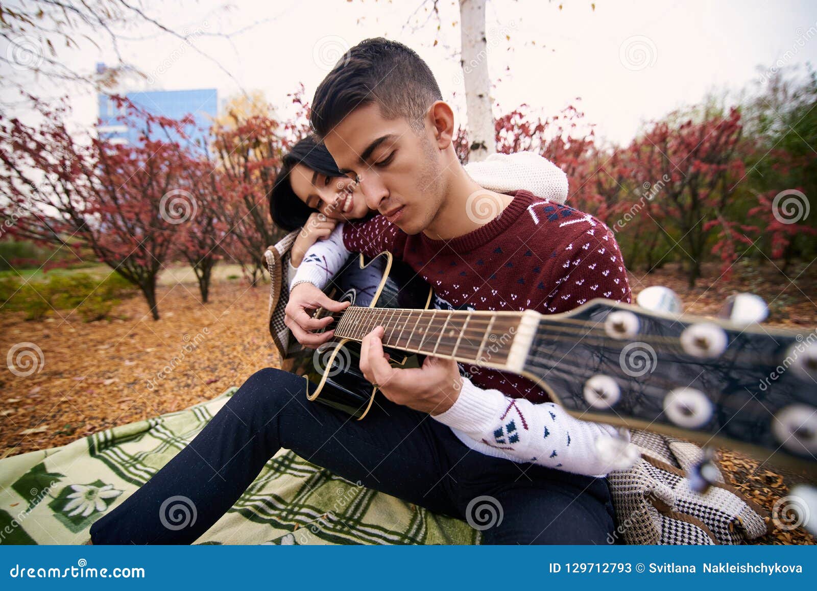 Individuo con una manta cubierta muchacha. El individuo de piel morena hermoso toca la guitarra, y al lado de él sienta a una muchacha hermosa que puso su cabeza en su hombro