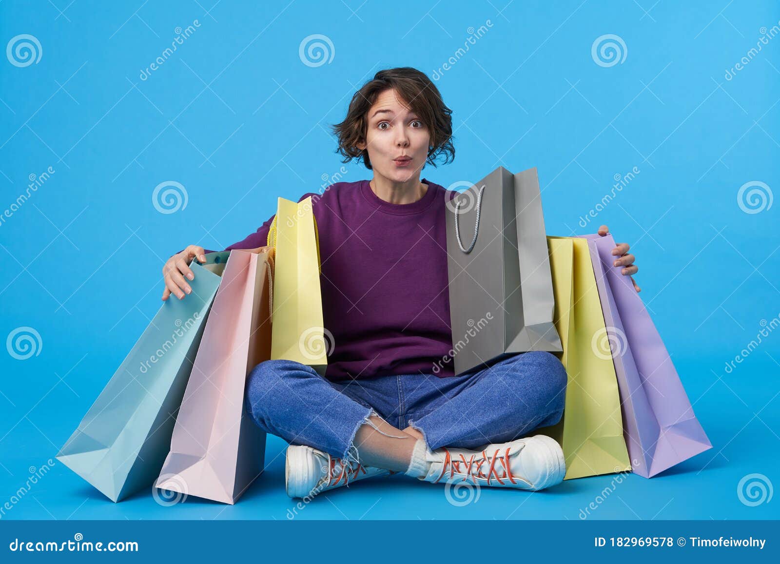 indoor photo of amazed young lovely short haired curly lady raising surprisedly eyebrows while looking at camera, sitting over