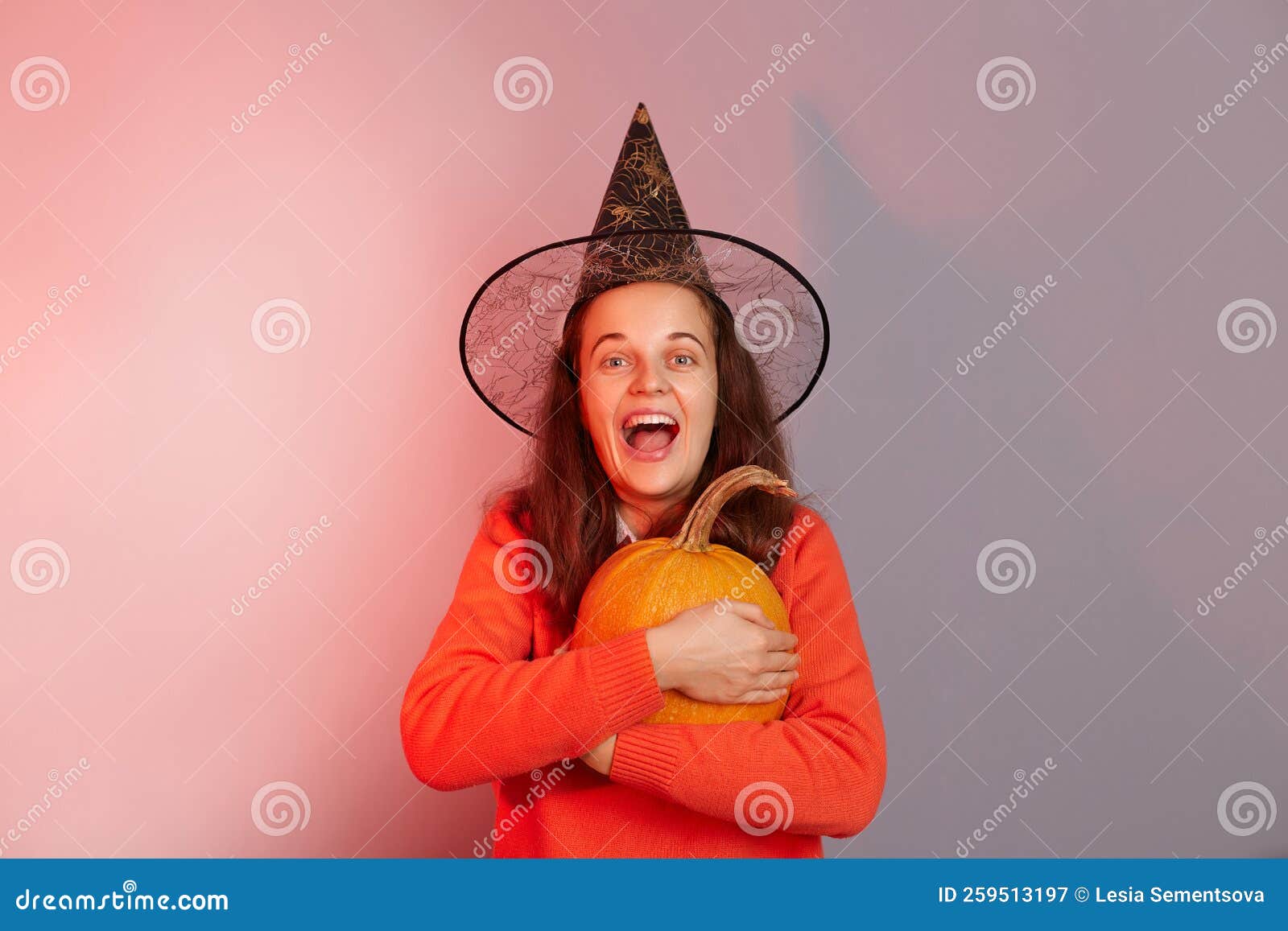 Indoor Shot Of Excited Amazed Caucasian Woman Dressed In Witch Hat Holding Orange Pumpkin