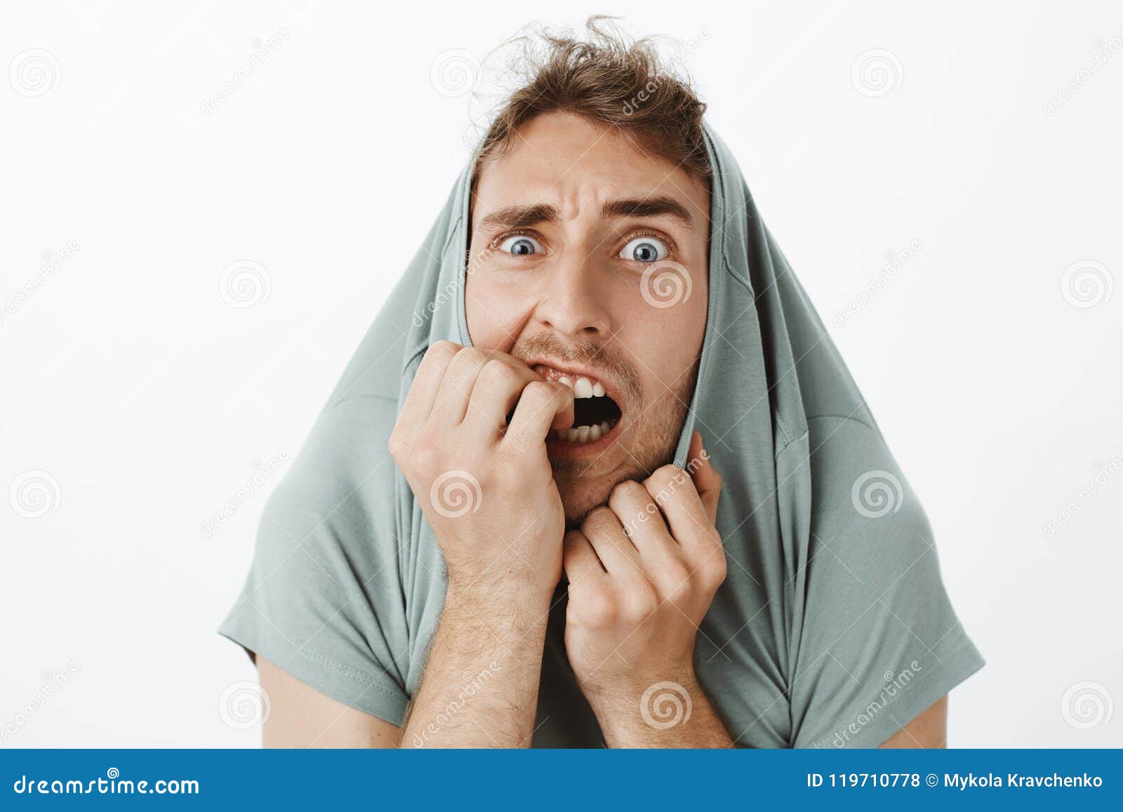 indoor shot of funny scared caucasian guy with bristle, pulling t-shirt on head and staring with popped eyes through