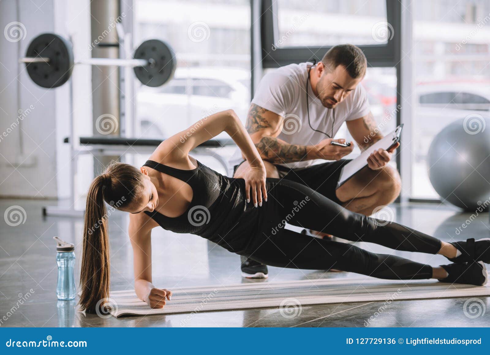 Instructor personal masculino que mira el contador de tiempo y a la mujer atlética joven que hacen el tablón lateral en la estera de la aptitud