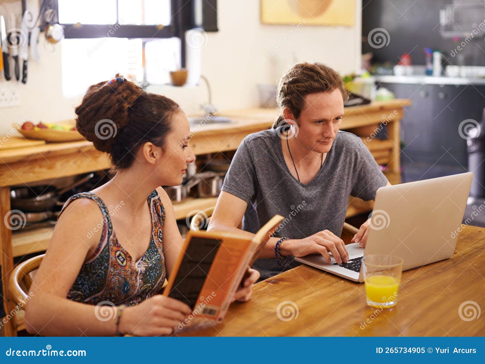 internet trumps book every time. a happy couple with dreadlocks sitting at a table using a laptop and reading a book.