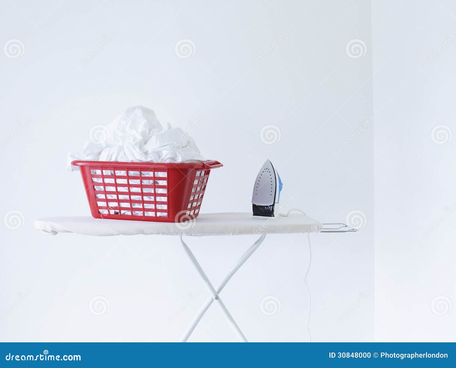 Iron and Laundry Basket on Ironing Board Against White Wall Stock Photo