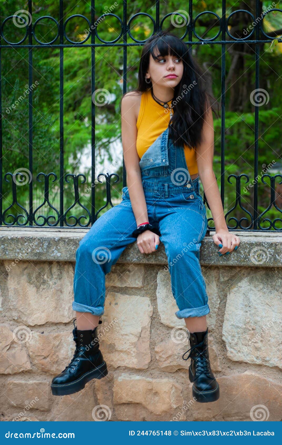 Jeune Fille Habillee En Jaune Et Portant Une Combinaison En Denim Assise Sur Un Mur En Pierre Dans Un Parc Aux Couleurs Vert Et Ja Photo stock Image du languettes denim