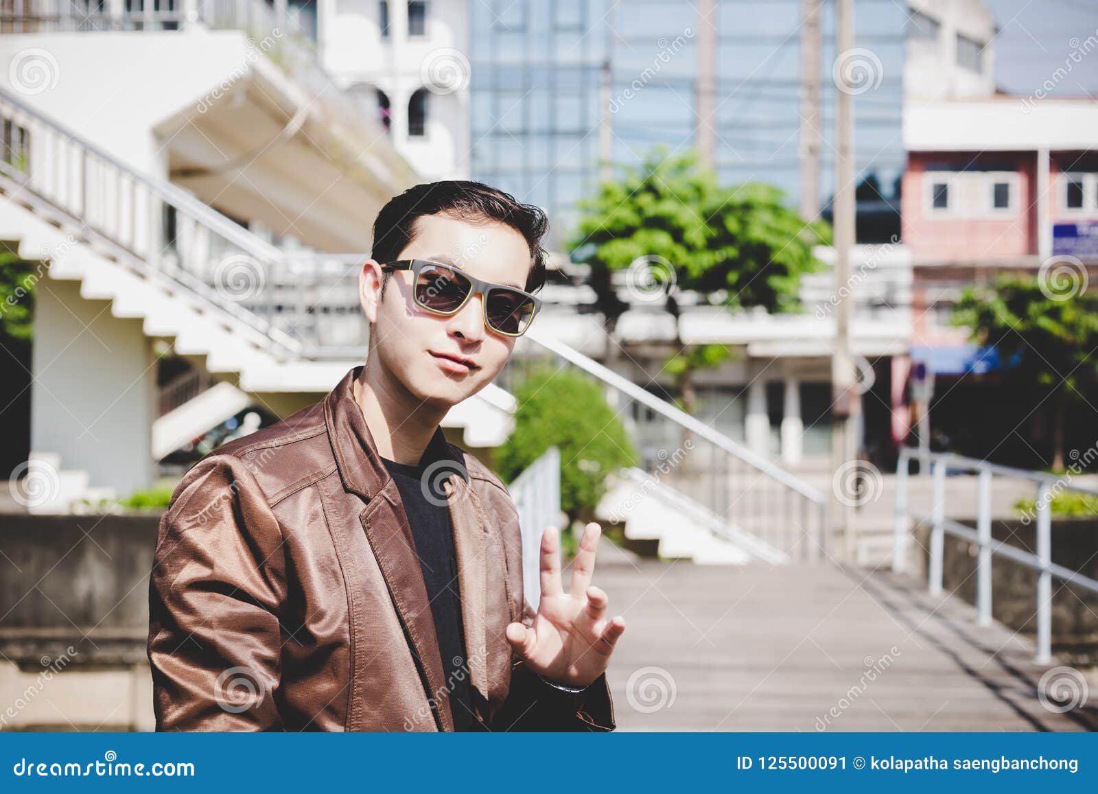 Jeune homme beau avec du charme de portrait Le type attirant regarde l'ami ou quelqu'un et ondule sa main pour dire le bonjour tandis qu'il marche dans la ville L'homme de charme utilise des lunettes de soleil, veste en cuir