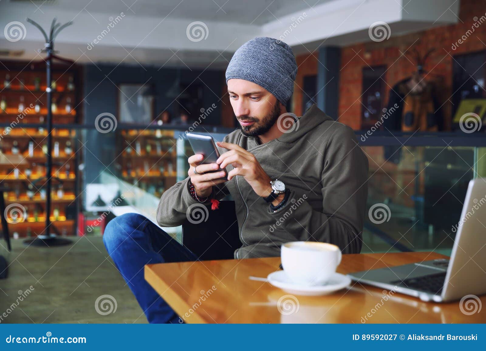 Jeune homme bel de hippie avec la barbe se reposant dans le téléphone portable parlant de café, tenant la tasse de café et de sou. Jeune homme bel de hippie avec la barbe se reposant dans le téléphone portable parlant de café, tenant la tasse de café table d'ordinateur portatif en bois