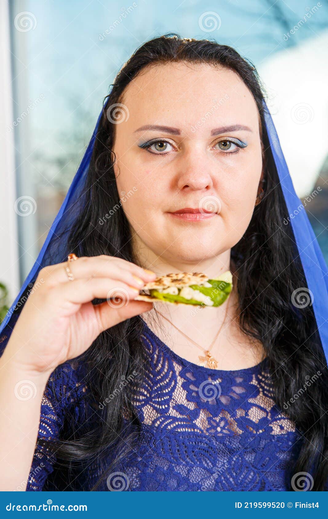 a jewish woman with her head covered in a blue cape at the passover seder table is eating moror hazeret matzah.