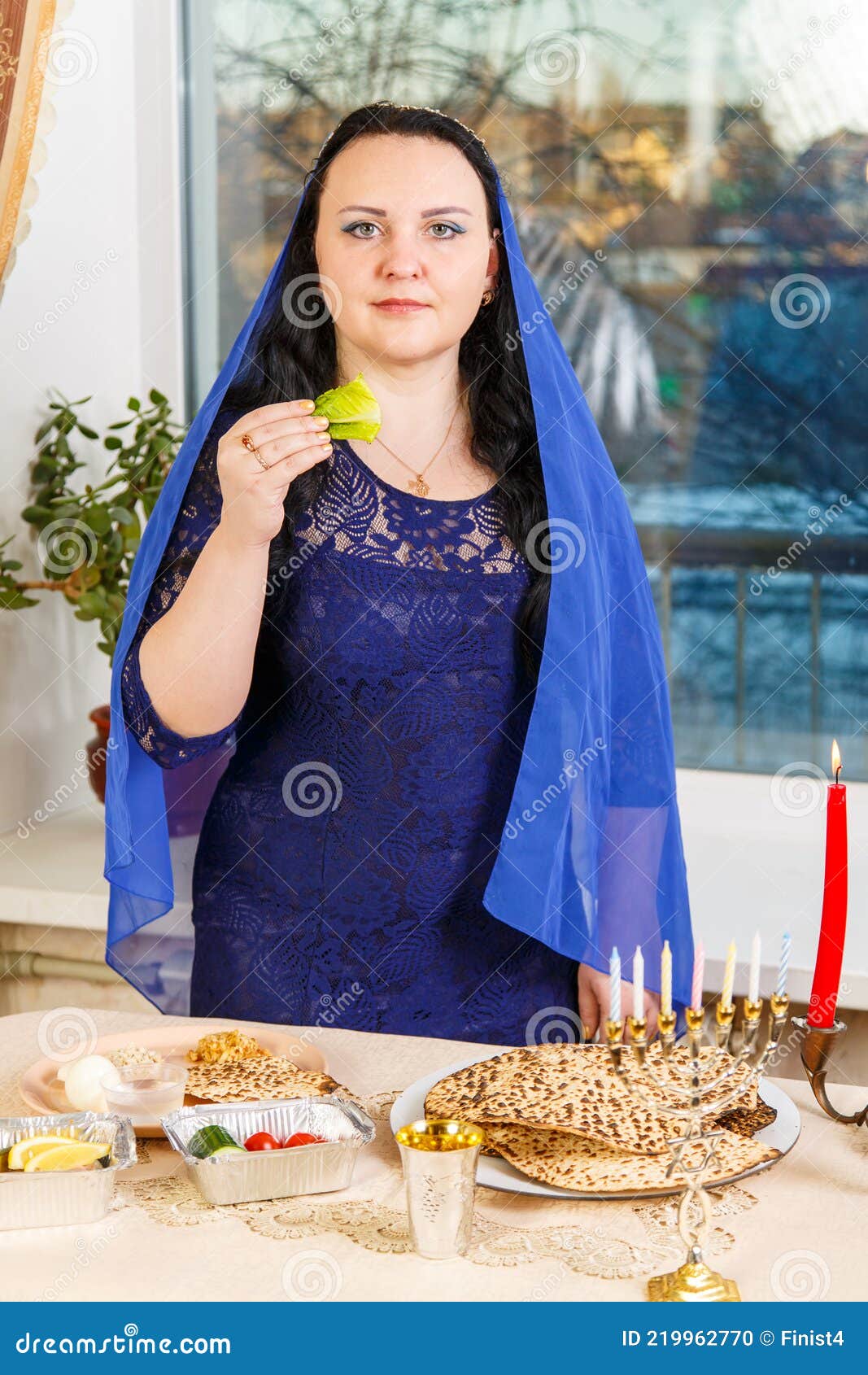 a jewish woman with her head covered in a blue cape at the passover seder table is eating moror hazeret matzah.