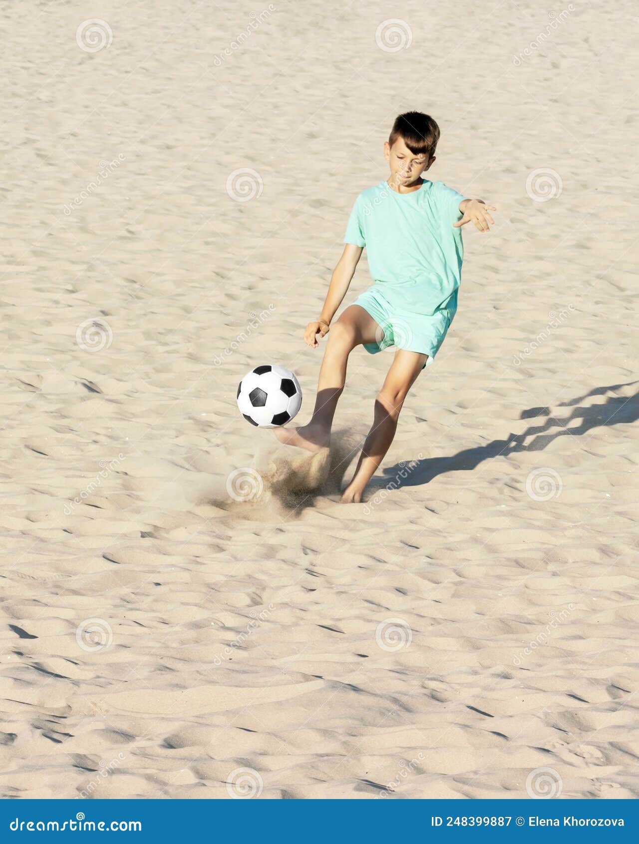 Garoto Joga Bola De Futebol Na Praia Arenosa. Pés Descalços De