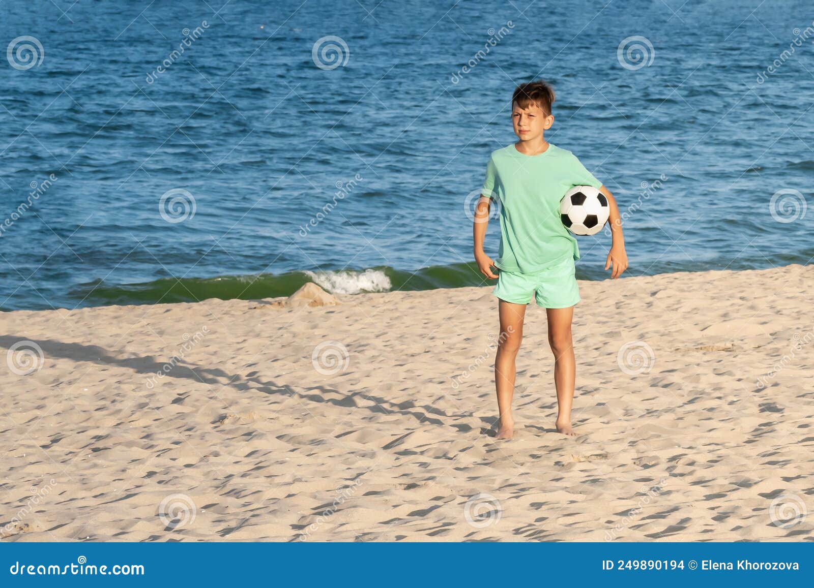 Garoto Joga Bola De Futebol Na Praia Arenosa. Pés Descalços De