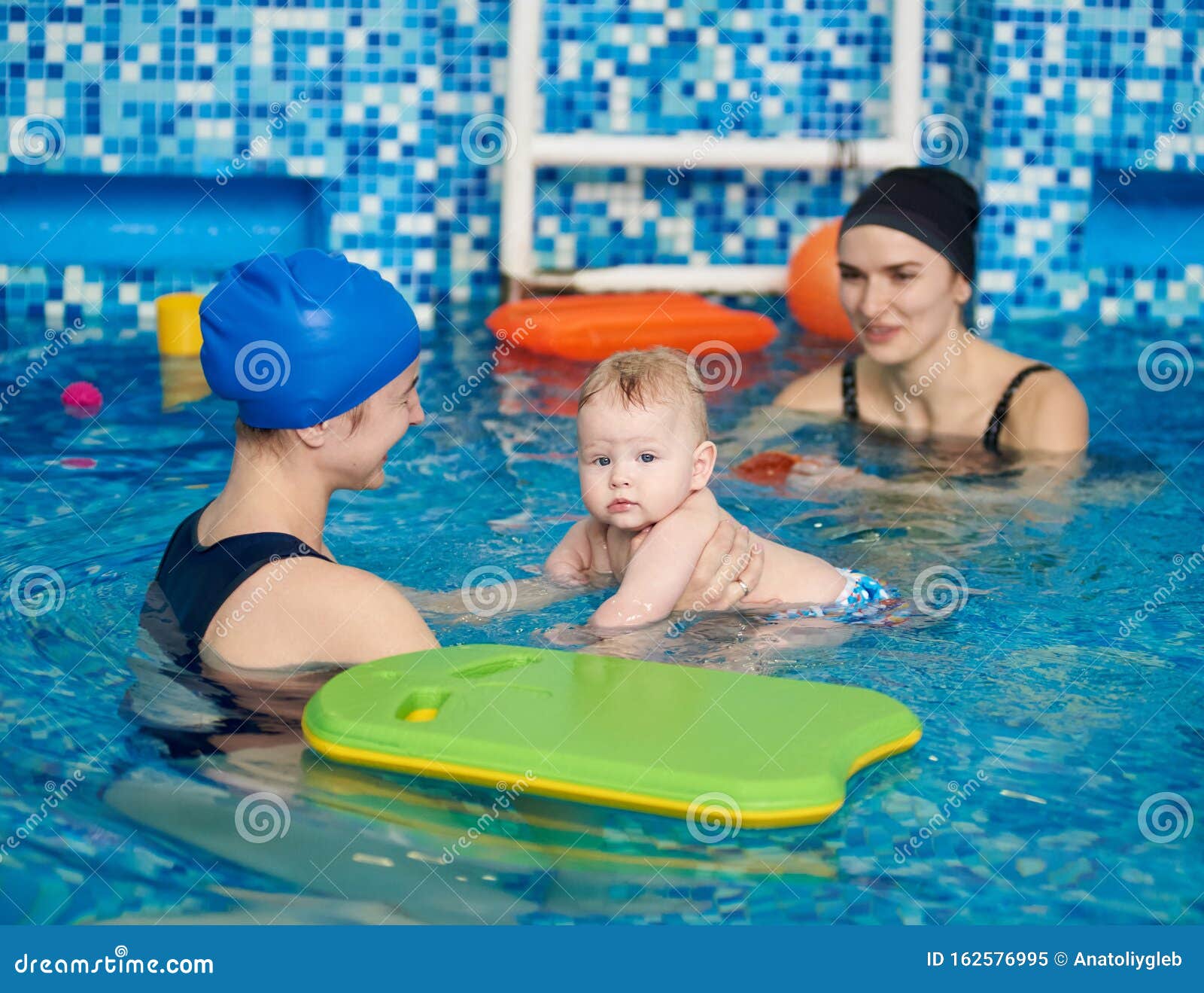 Jonge gelukkige moeder en baby genieten van zwevend zwemmen terwijl ze opleiders in het zwembad oplichten. Zijaanzicht van jonge glimlachende moeder en haar baby genieten van het paddling zwemmen met trainer in het zwembad Kind in de handen van moeder, opzij kijkend naar het zwembad Begrip geluk en liefde