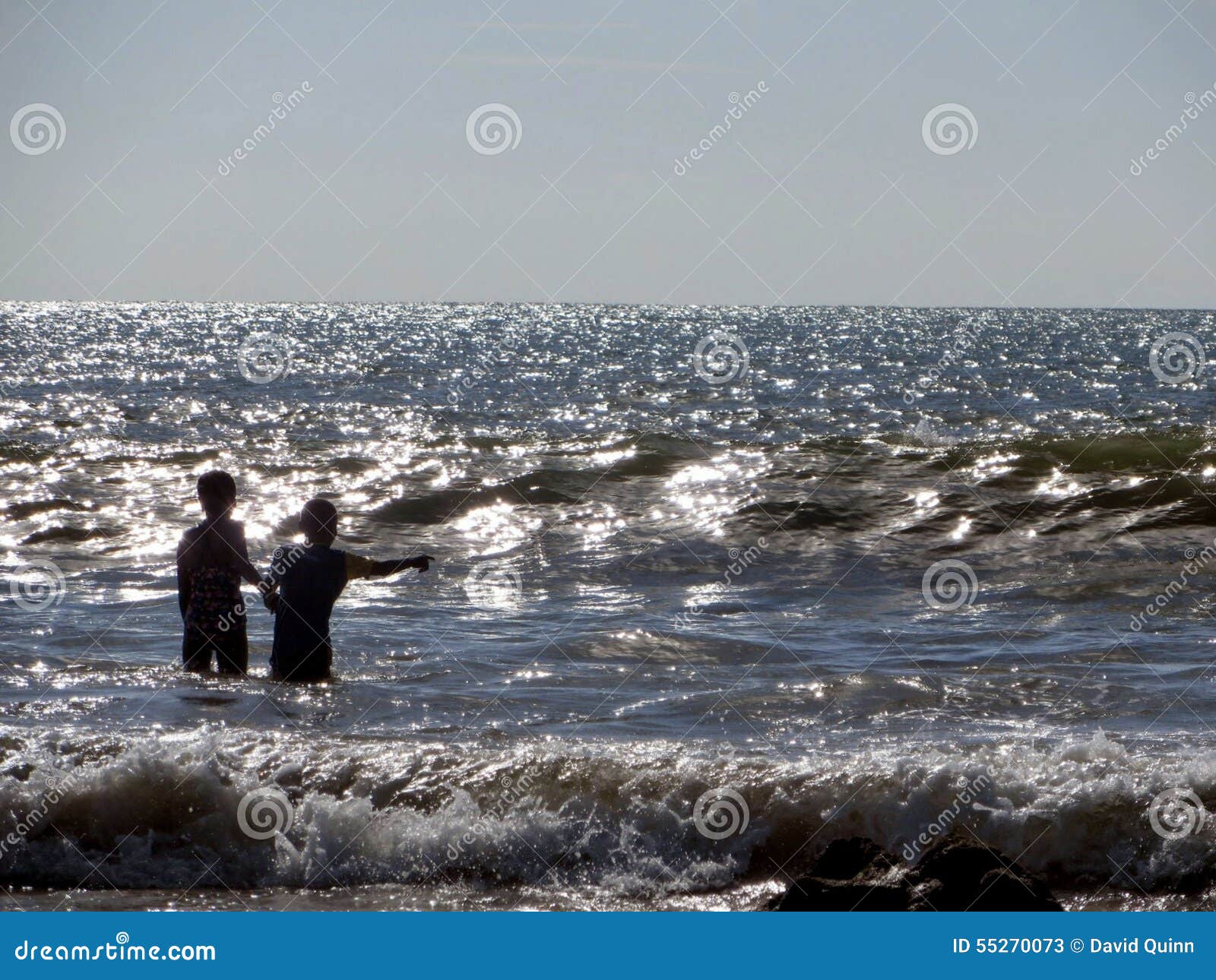 Jonge kinderen bij spel in de Indische Oceaan van de kust van Koh Lanta Thailand. Een jong jongen en een meisje bevinden zich in de glorierijke Indische Oceaan van de kust van Ko Lanta Thailand Als zonvlekken bevindt het water zich, het silhouet van de kinderen in schril contrast aan het blauw van het water en grijs van de hemel De golven blijven tegen het strand aan het front van de foto verpletteren