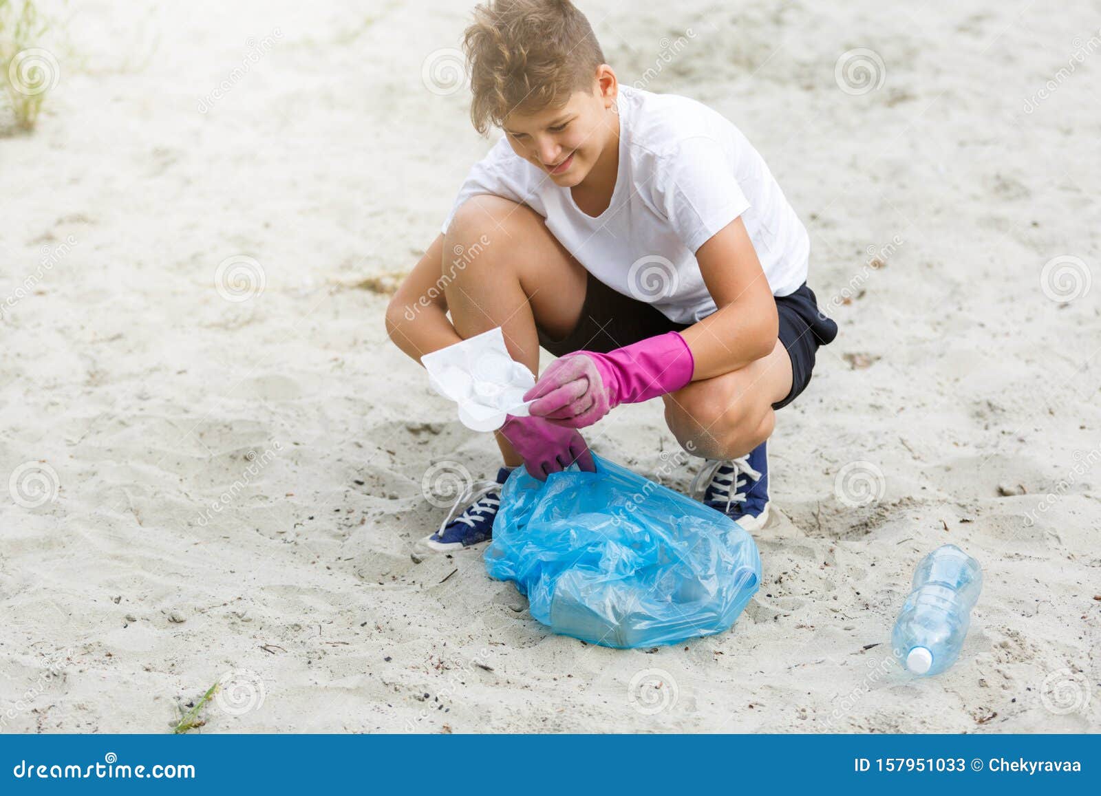 Jongen in wit T-shirt in handschoenen verzamelt vuilnis en plastic flessen in blauwe verpakking op het strand Jonge vrijwilliger Milieubescherming, begrip "sparen milieu" Handen in handschoenen sluiten