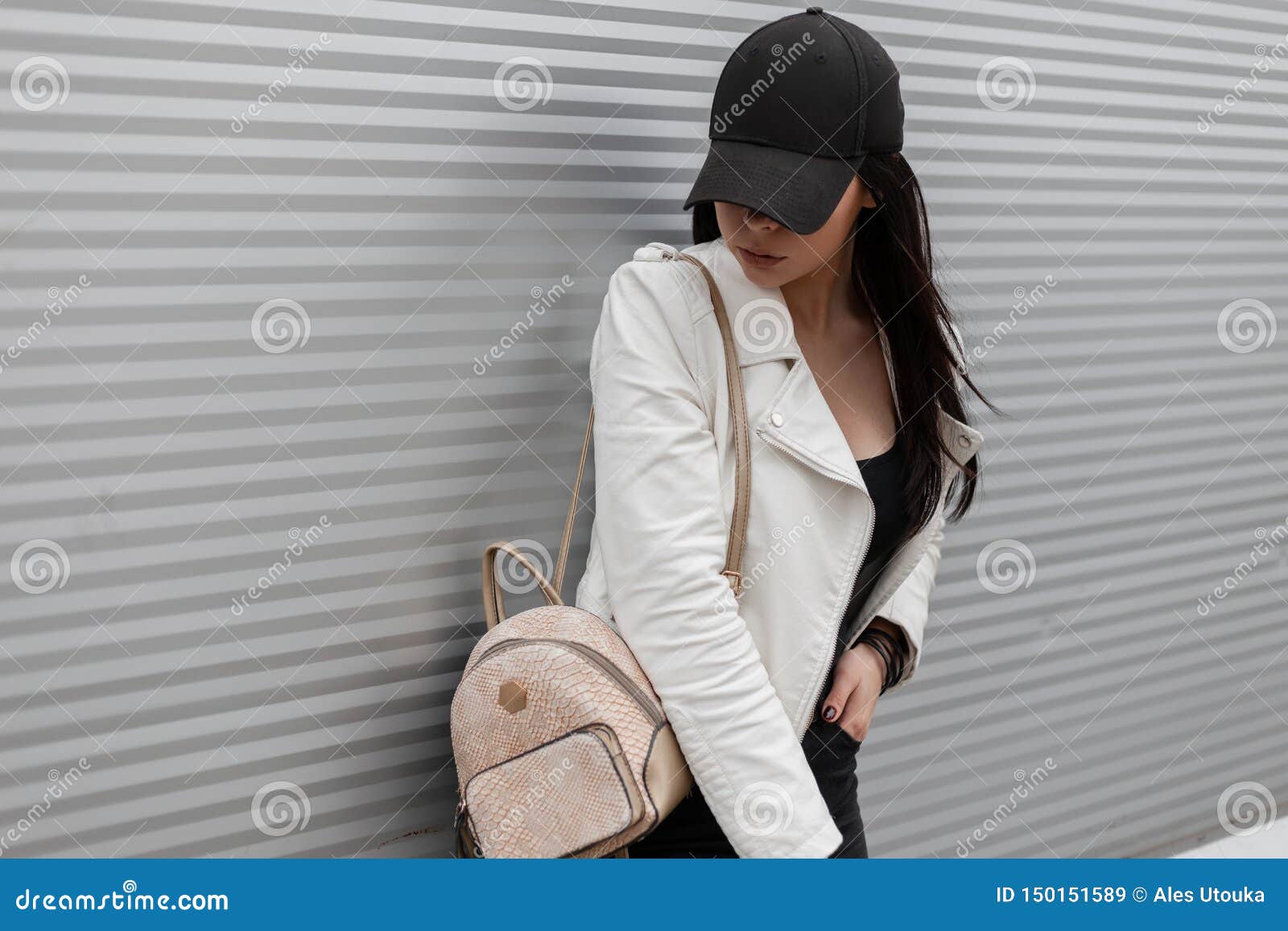 A jovem mulher moderna bonita em um boné de beisebol preto elegante em um casaco de cabedal branco do vintage nas calças de brim com uma trouxa do ouro está estando perto de uma parede do metal e está olhando para baixo Ar livre americano da menina